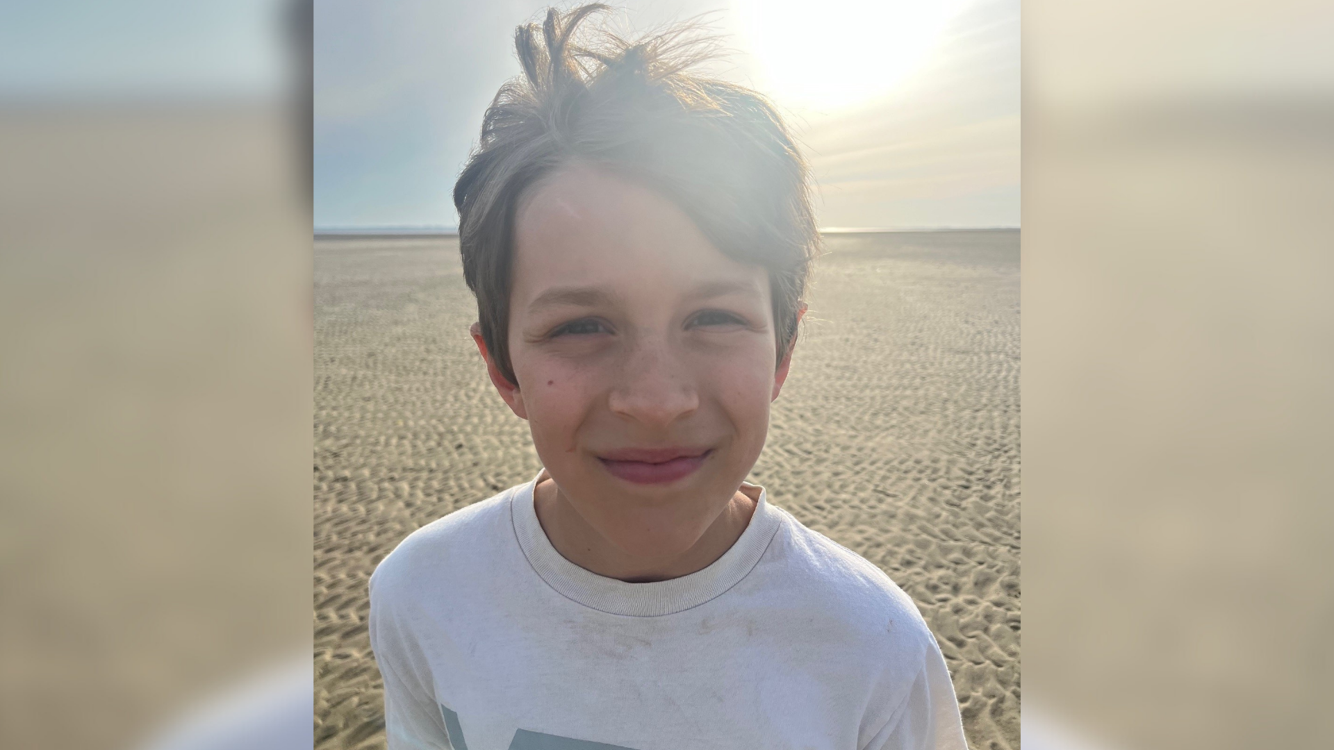 Ein Junge steht am Strand und lacht in die Kamera. 