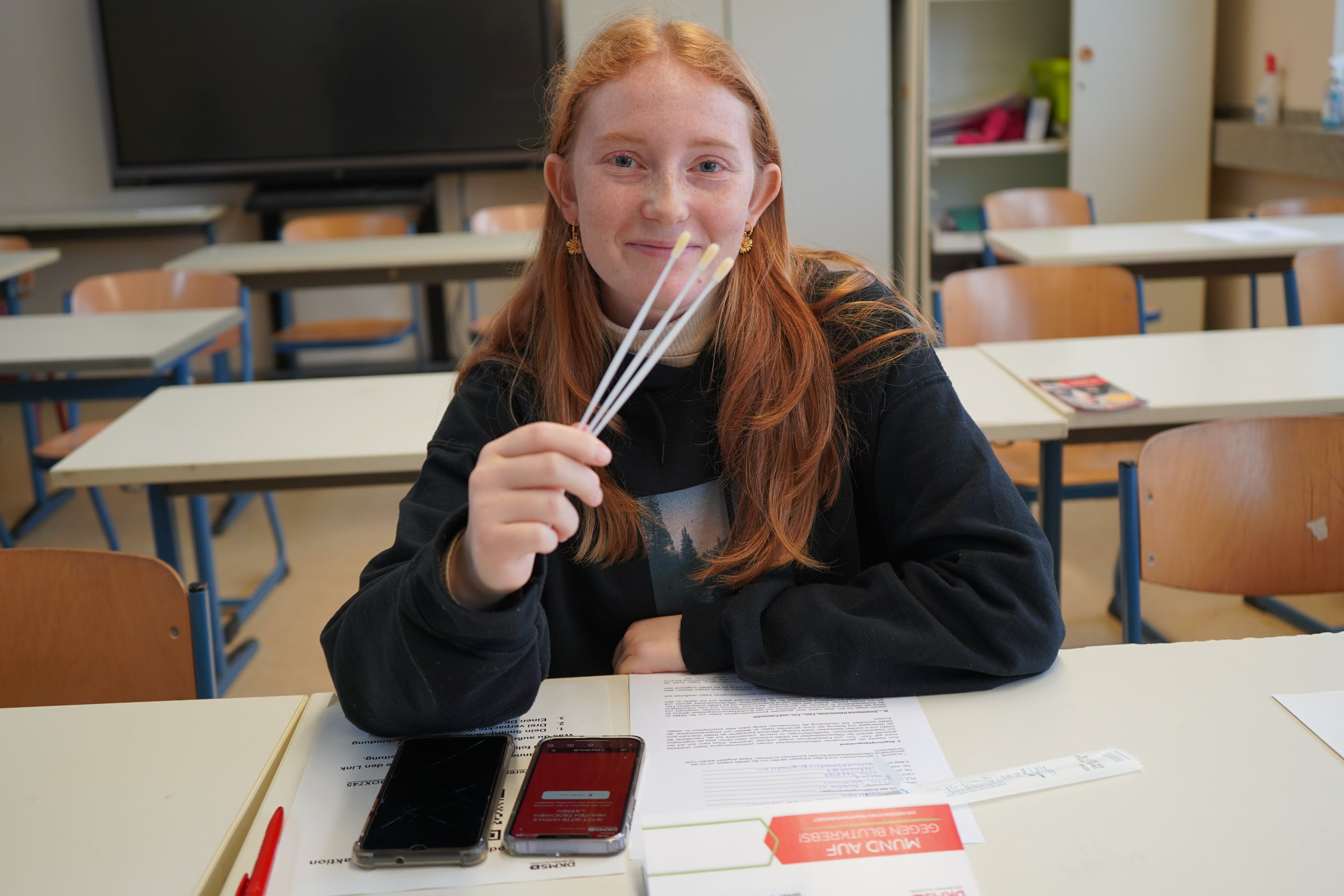 Junge Frau mit roten Haaren sitzt in einem Klassenzimmer an einem Tisch und hält drei Wattestäbchen in der Hand. Vor ihr liegen Formulare, zwei Handys und eine DKMS-Spenderkarte. Sie lächelt in die Kamera.