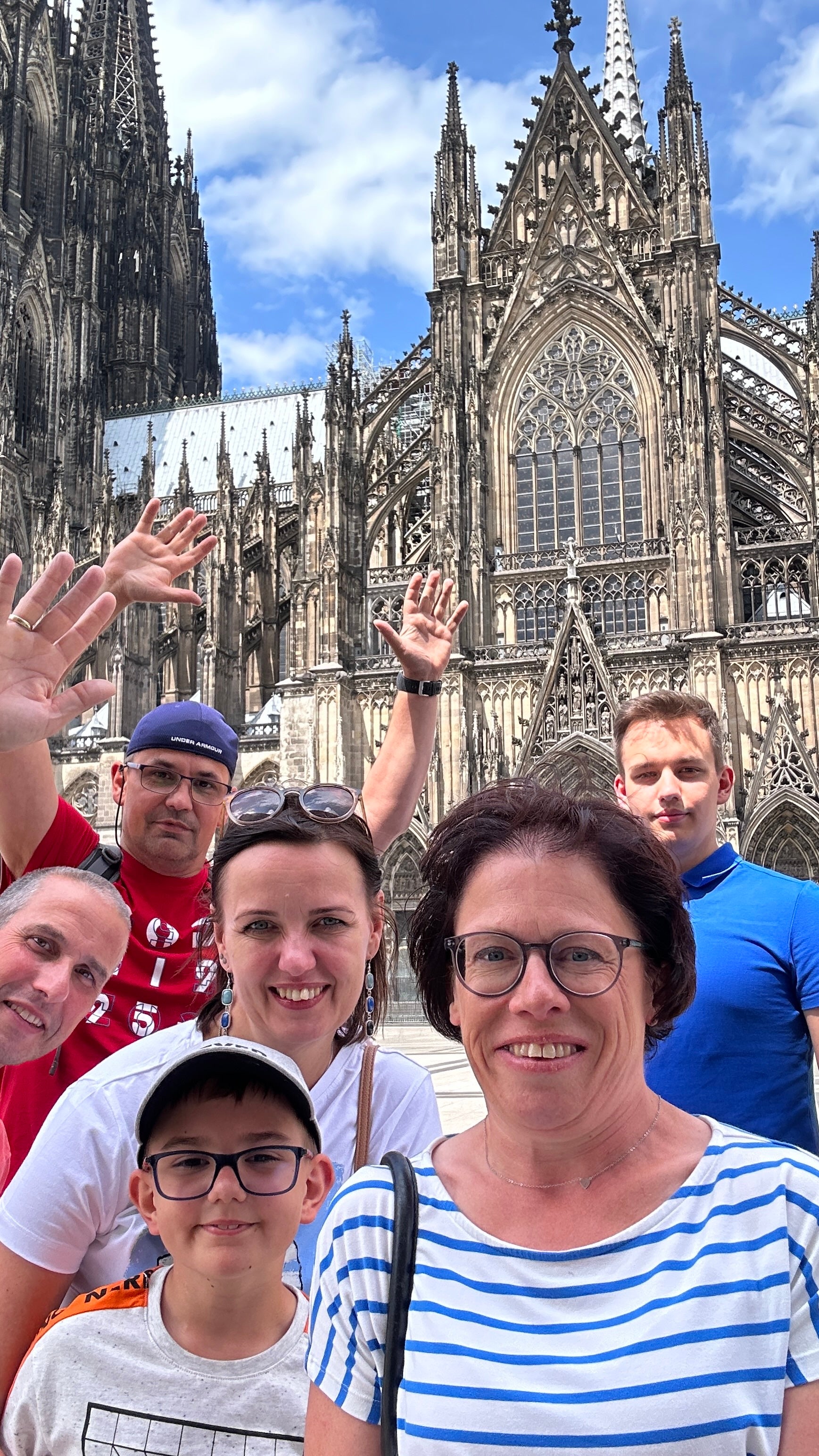 Sieben Personen stehen eng zusammen vor dem Kölner Dom und blicken in die Kamera. Einige winken, andere lächeln. Im Vordergrund stehen zwei Frauen und ein Junge mit Brille, im Hintergrund sind Erwachsene in farbigen T-Shirts zu sehen. Der Kölner Dom mit seinen markanten Türmen und Fassadendetails füllt den Hintergrund. 