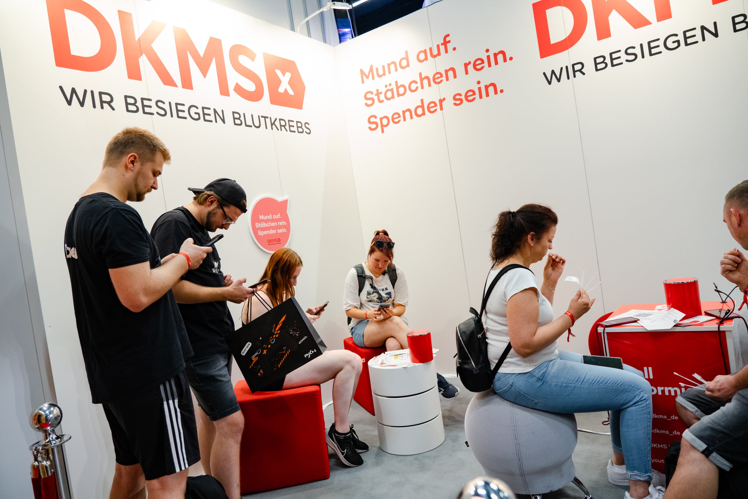 Mehrere Personen sitzen und stehen an einem DKMS-Messestand und füllen Registrierungsunterlagen aus. Der Hintergrund zeigt die DKMS-Wand mit dem Schriftzug „Mund auf. Stäbchen rein. Spender sein.“ und „Wir besiegen Blutkrebs“. Einige Personen halten Wattestäbchen oder Formulare in den Händen. Die Atmosphäre ist ruhig und konzentriert.
