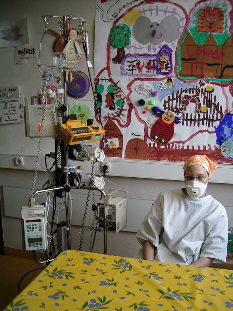 Stella sitzt in einem Krankenhaus. Sie trägt ein weißes Gewand und eine Atemschutzmaske.