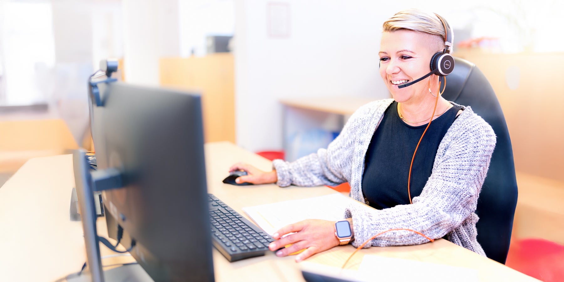 Eine blonde Frau, die ein Headset trägt und an einem Schreibtisch sitzt und telefoniert