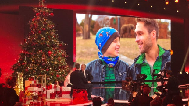 Eine Bühne, auf der ein großer Weihnachtsbaum steht. Im Hintergrund ein Bildschirm, auf der man einen Mann und einen Jungen sieht