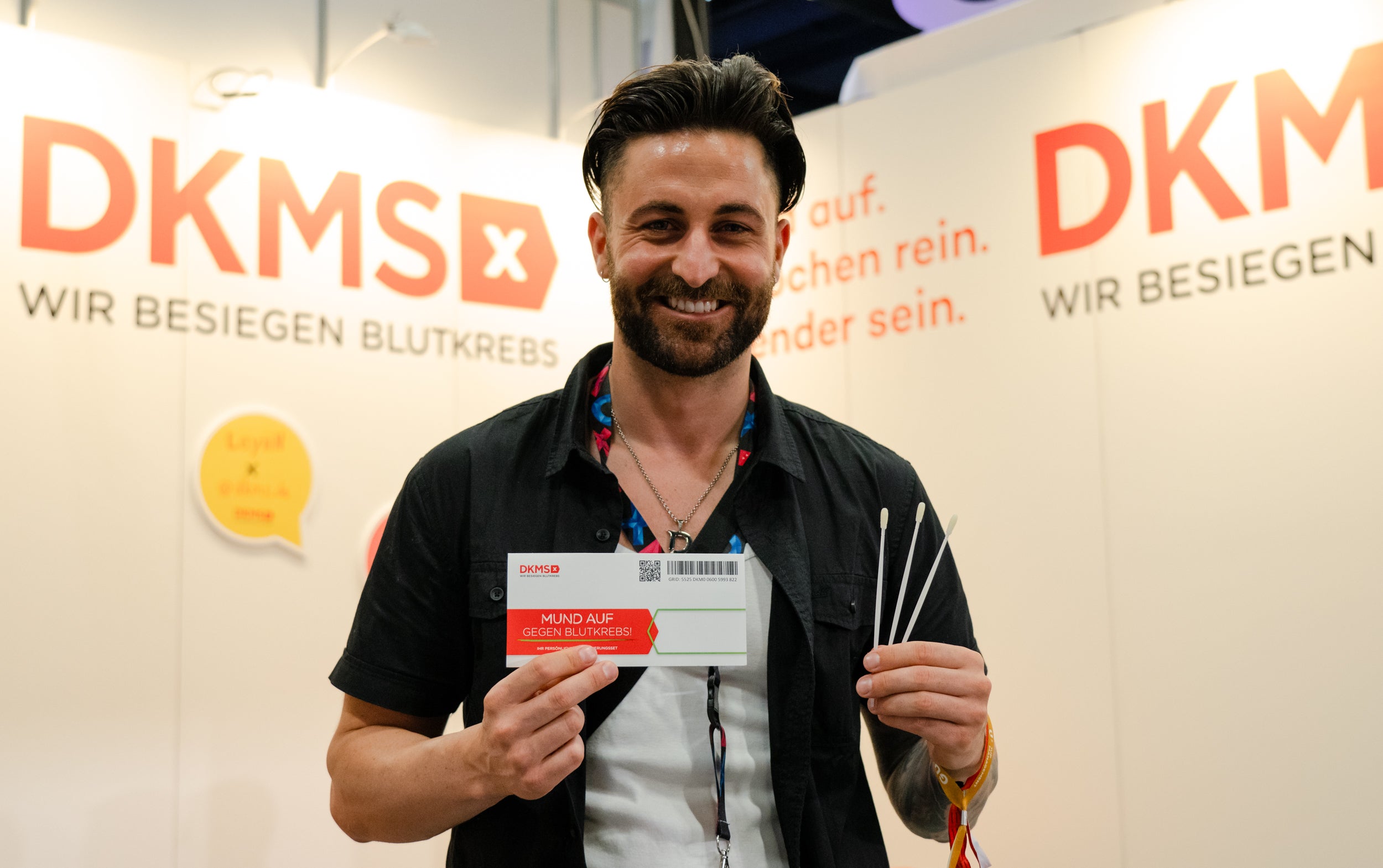 Ein Mann steht vor einem DKMS-Messestand mit der Aufschrift „Wir besiegen Blutkrebs“. Er lächelt in die Kamera und hält ein DKMS-Registrierungsset sowie mehrere Wattestäbchen in den Händen. Im Hintergrund sind Sprechblasen-Grafiken und ein hell beleuchteter Messestand mit Textausschnitten wie „Mund auf. Stäbchen rein. Spender sein.“ zu sehen.
