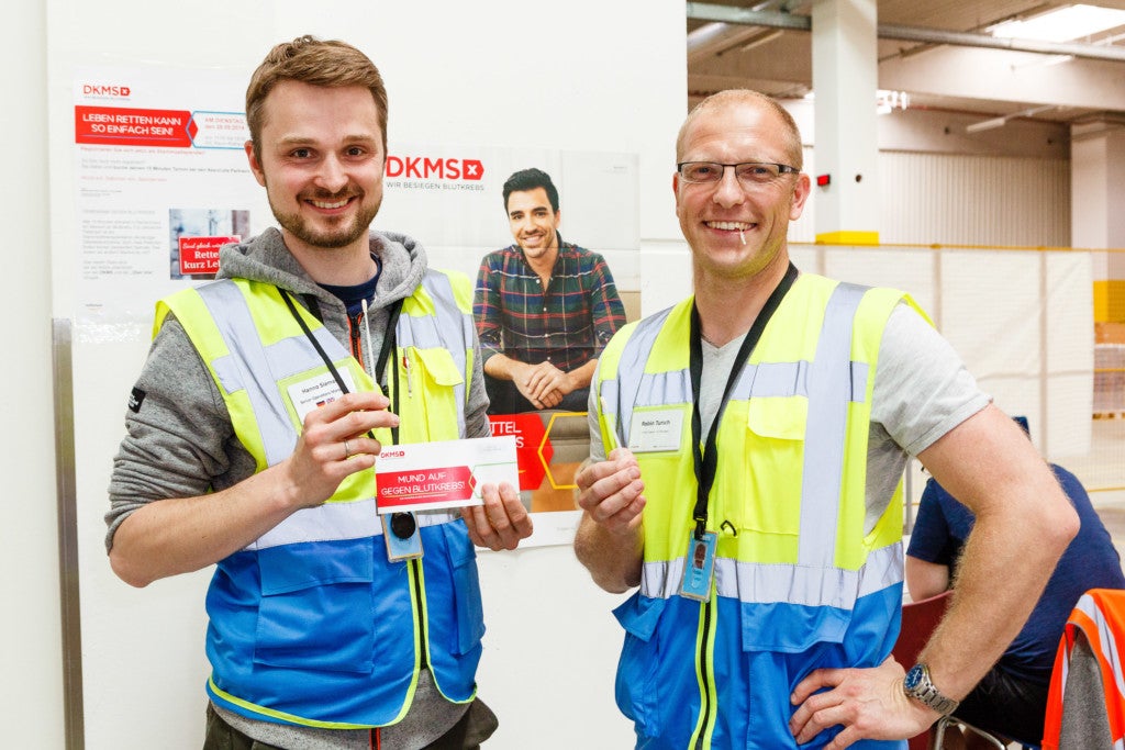 Zwei Männer, die reflektierende Westen tragen und in einem Raum stehen. Der Mann links hält ein Registrierungset der DKMS