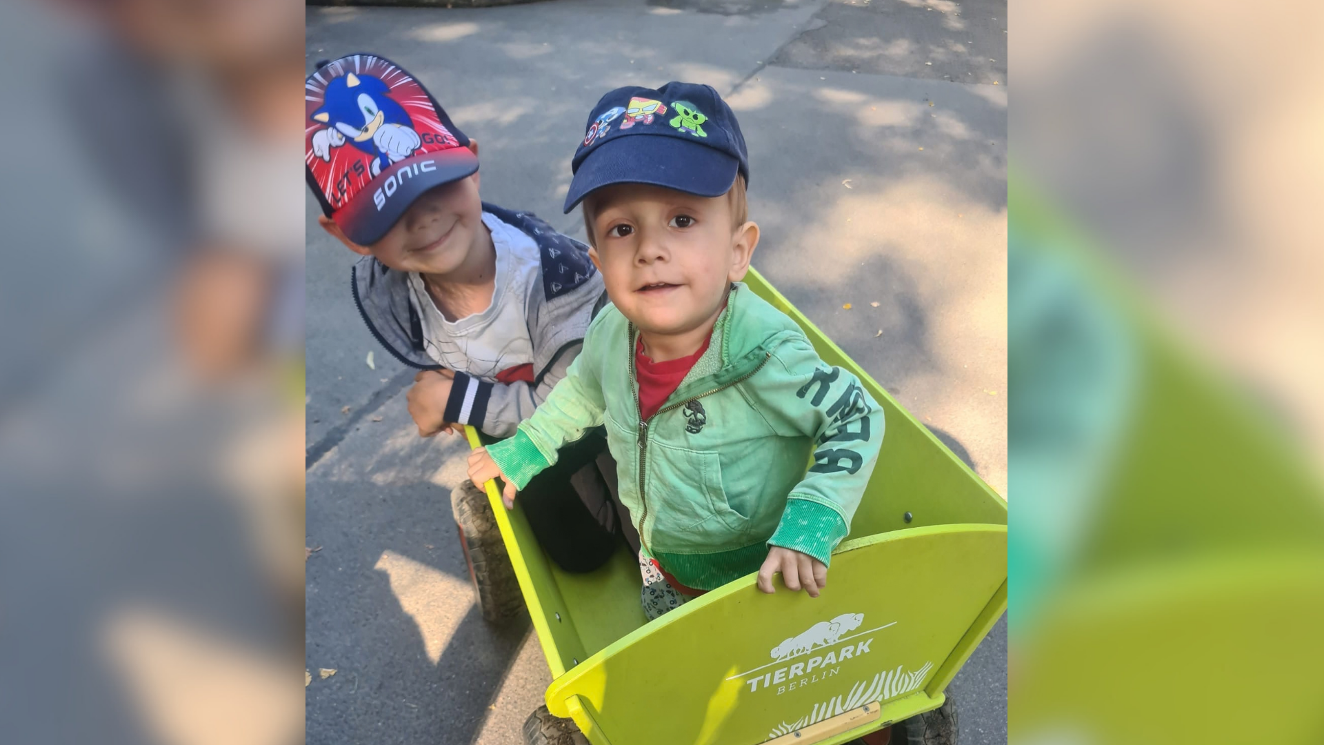 Zwei kleine Jungen tragen Kappen und sitzen in einem grünen Bollerwagen. 