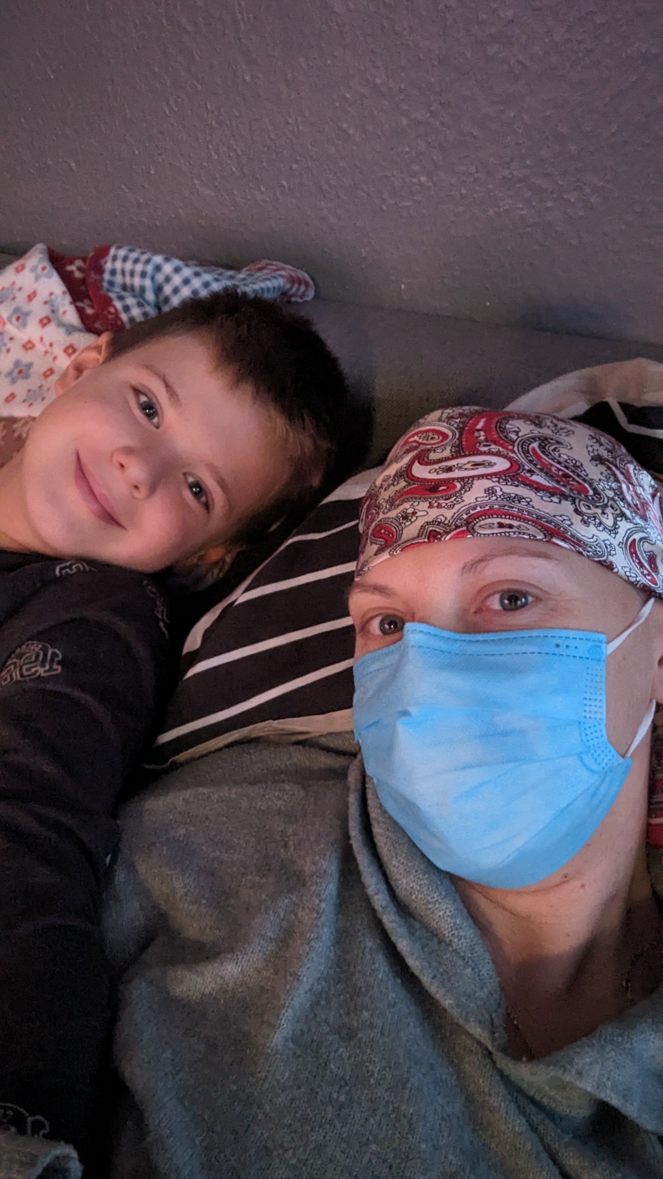 Ein Kind und eine erwachsene Person liegen nebeneinander und schauen in die Kamera. Das Kind lächelt, die erwachsene Person trägt ein Kopftuch mit Muster und eine medizinische Maske. Beide liegen auf gestreiften Kissen, der Hintergrund ist eine graue Wand.