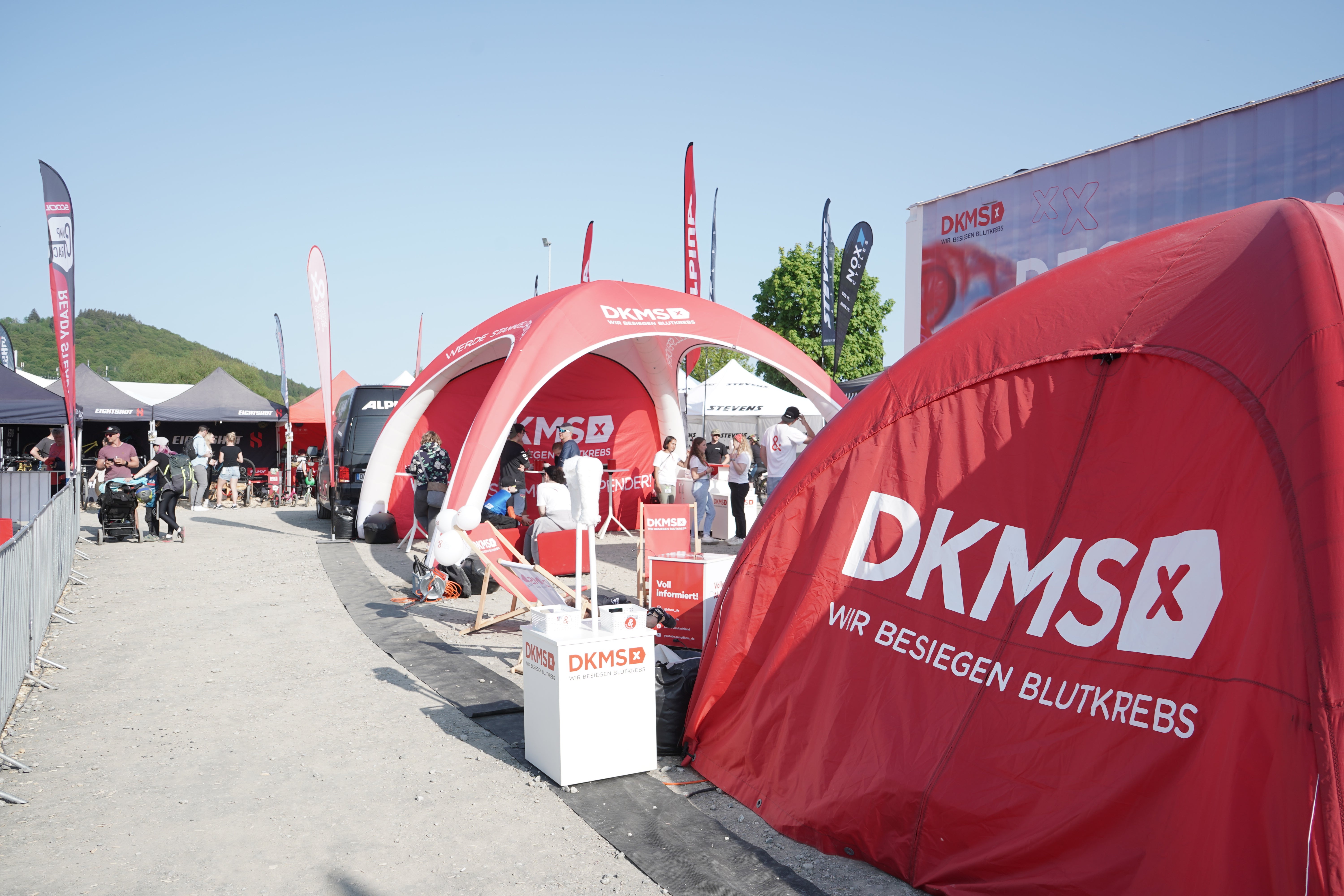 Rote Zelte und Pavillons mit DKMS-Logo stehen auf einem Veranstaltungsplatz im Freien. Auf den Zelten ist der Schriftzug „DKMS – Wir besiegen Blutkrebs“ zu sehen. Im Hintergrund befinden sich weitere Stände, Personen und Fahnen. Der Himmel ist blau und wolkenlos.