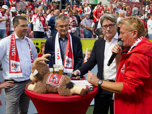 Eine Gruppe von Männern steht um einen Tisch mit einem Ziegen Kuscheltier 