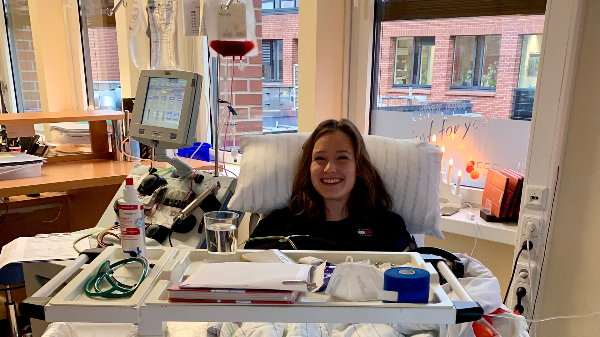 Eine junge Frau liegt lächelnd in einem Behandlungsstuhl während einer peripheren Stammzellentnahme. Vor ihr steht eine Apheresemaschine mit einem Infusionsbeutel, in dem sich Blutbestandteile befinden. Auf dem Rolltisch vor ihr liegen medizinische Utensilien und ein Glas Wasser. Im Hintergrund ist ein Fenster mit Blick auf ein Gebäude aus rotem Backstein sowie eine Kerze und eine Lichterkette auf der Fensterbank zu sehen.