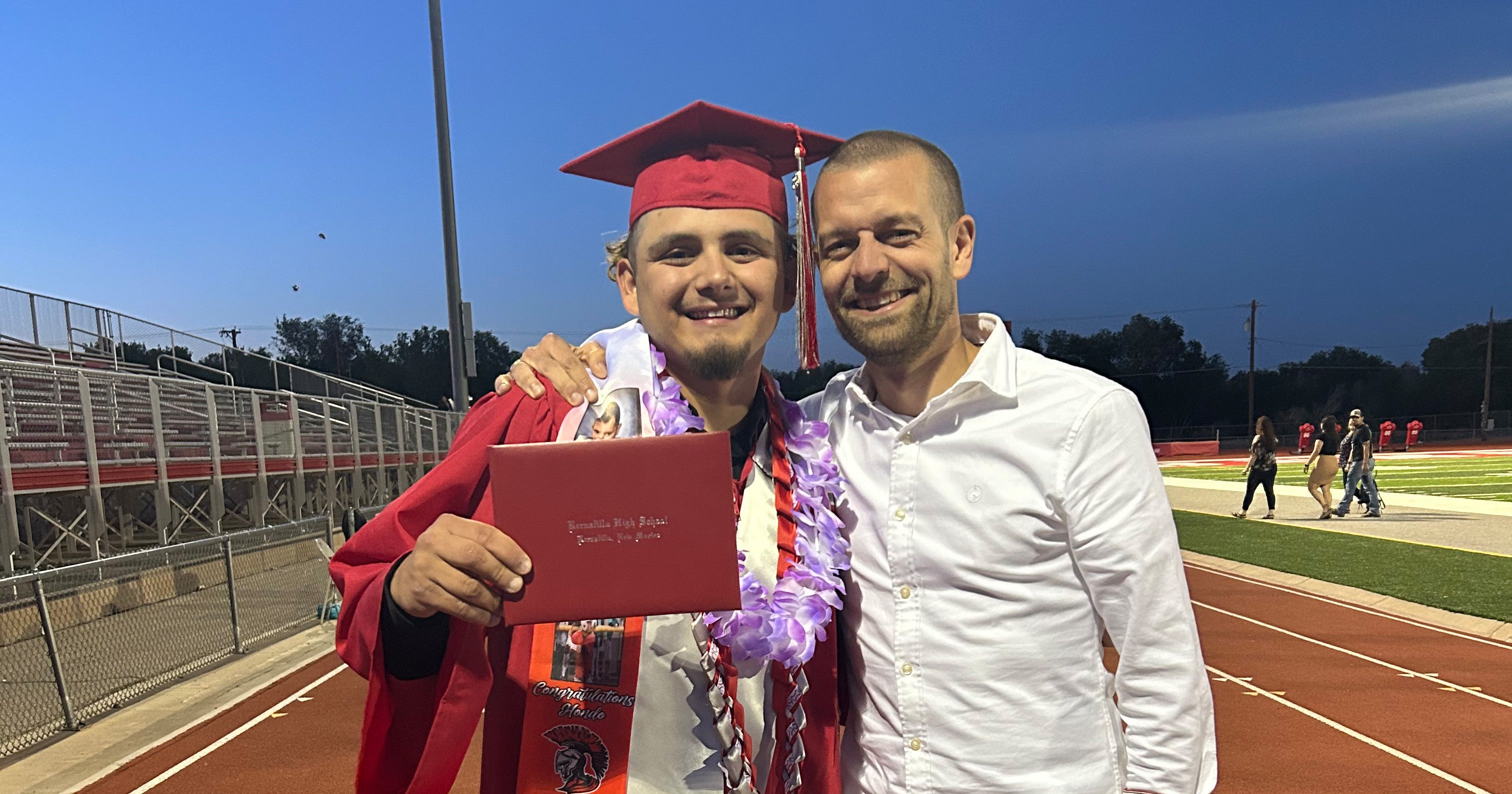 Zwei junge Männer lachen am Rande eine Sportfelds in die Kamera. Der eine trägt eine typische Robe zum Highschool-Abschluss.