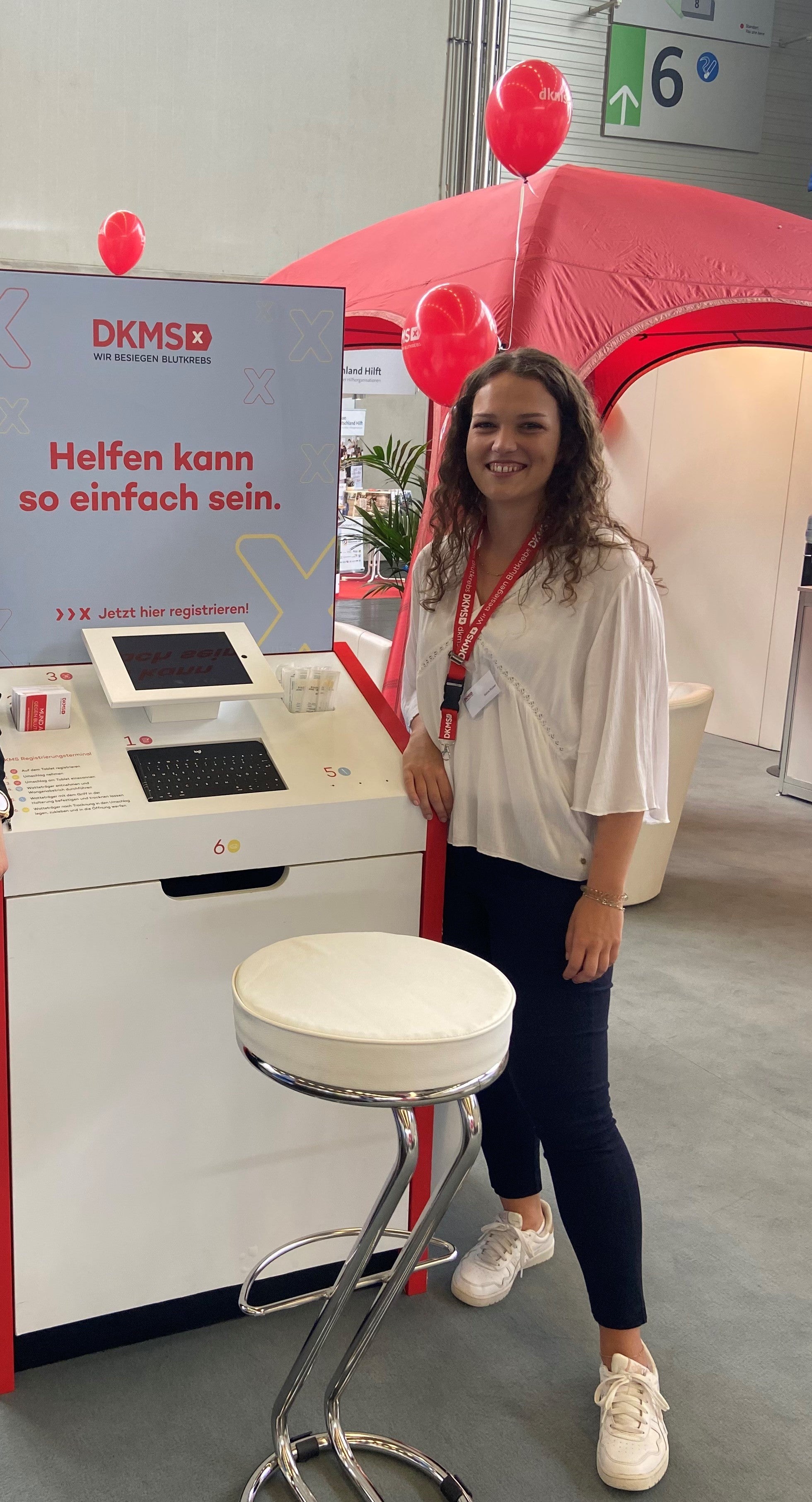 Eine Frau steht an einem DKMS-Informationsstand in einer Messehalle. Sie trägt ein weißes Oberteil, eine dunkelblaue Hose und weiße Turnschuhe. Um ihren Hals hängt ein Namensschild an einem roten Band. Auf dem Bildschirm am Stand steht „Helfen kann so einfach sein“. Im Hintergrund sind ein rotes Zelt, rote Luftballons und ein Schild mit der Hallenkennzeichnung „6“ zu sehen.