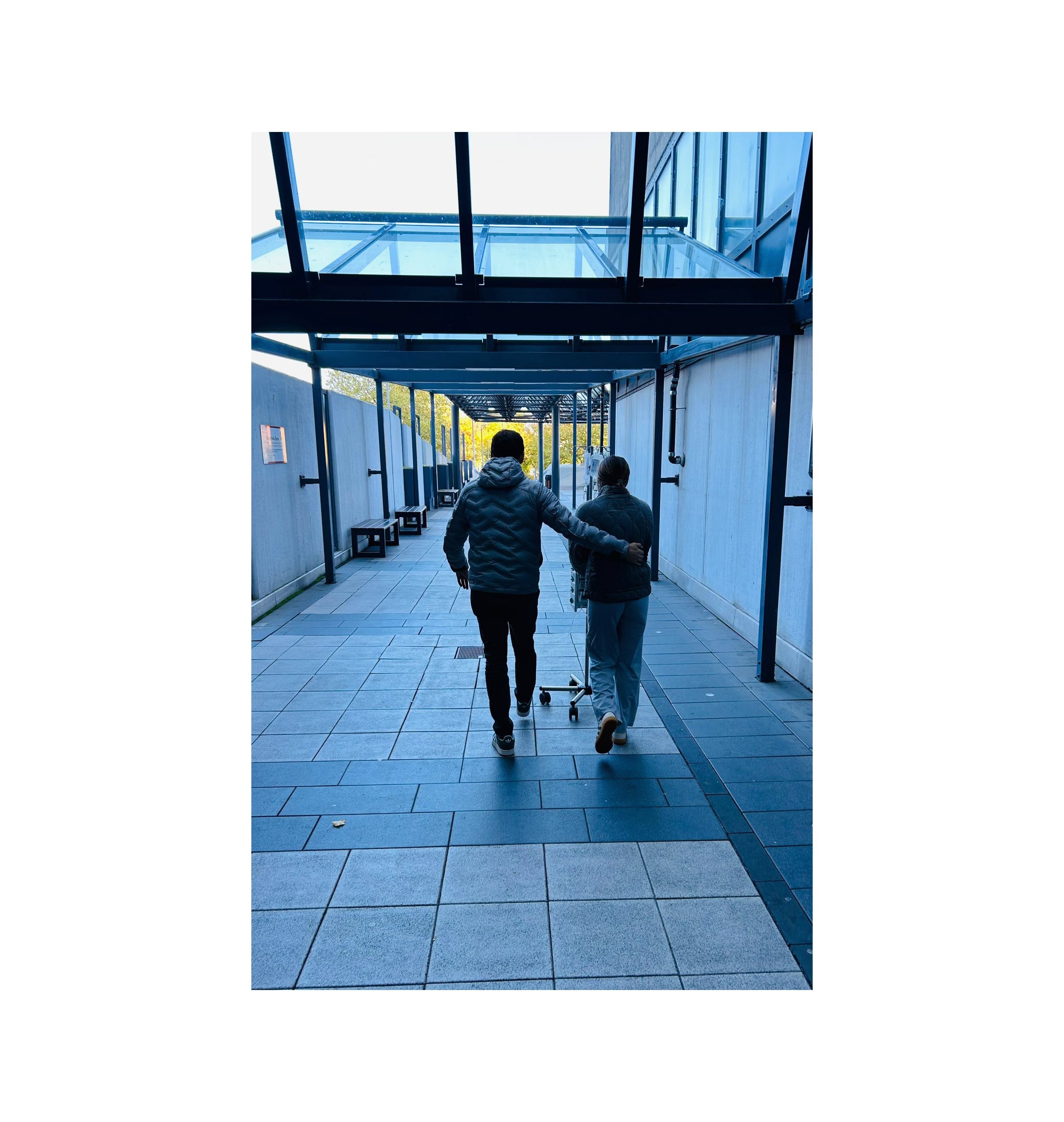 Das Bild zeigt zwei Personen von hinten, die gemeinsam durch einen überdachten Gang gehen. Eine Person hat den Arm um die andere Person gelegt. 