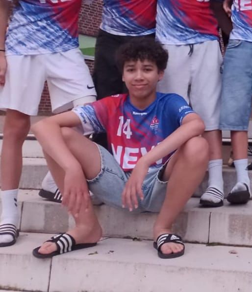 Ein Junge sitzt auf einer Treppenstufe. Er trägt ein blau-rotes Trikot und eine kurze Hose und um ihn herum sind weitere Menschen mit diesen Trikots zu sehen.