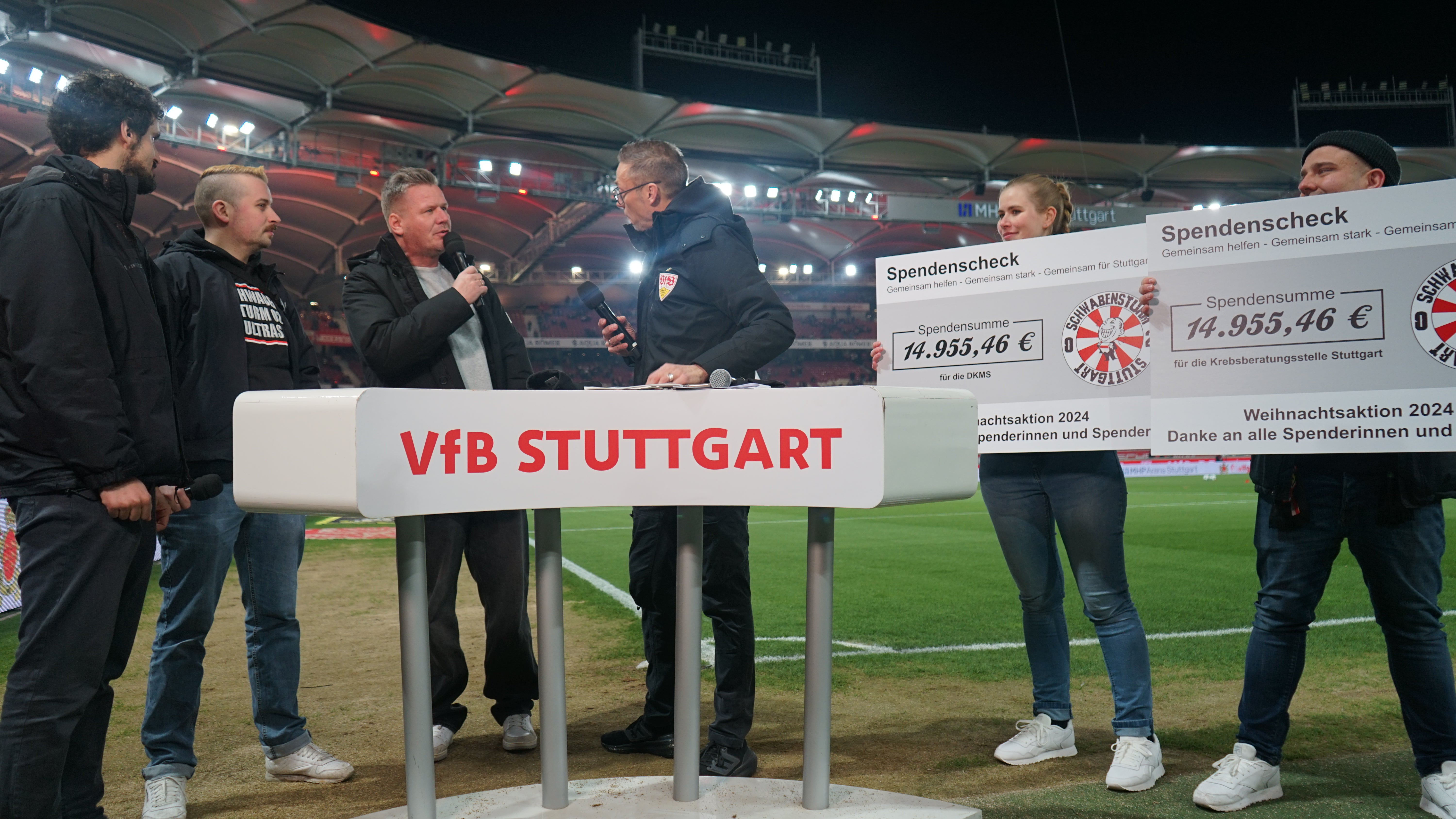 6 Menschen stehen am Rand von einem Fußballplatz. 2 Menschen halten einen Scheck hoch, die anderen 4 befinden sich in einer Interview Situation.