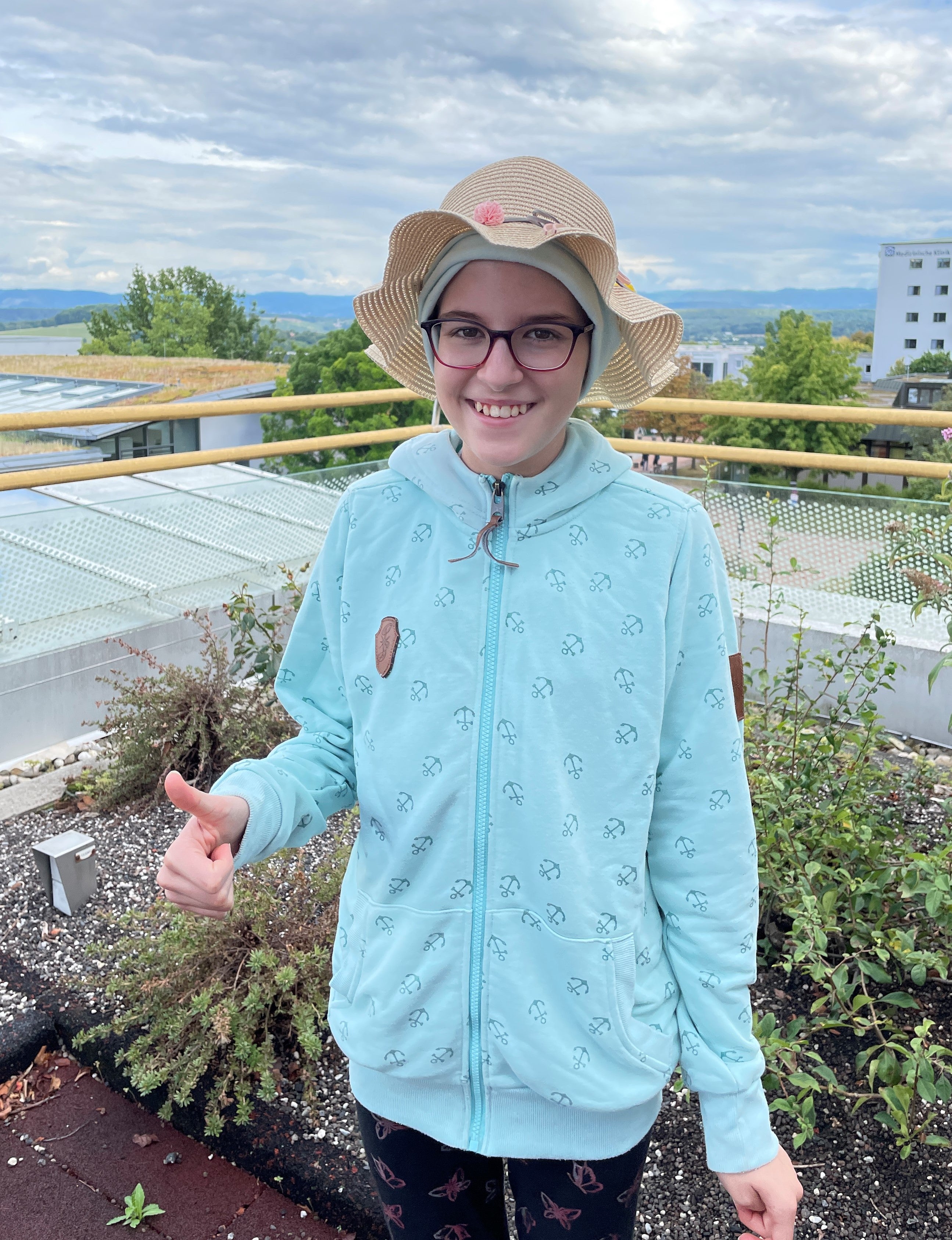 ein Kind steht mit Hut und Jacke auf einem Balkon und zeigt den Daumen hoch 