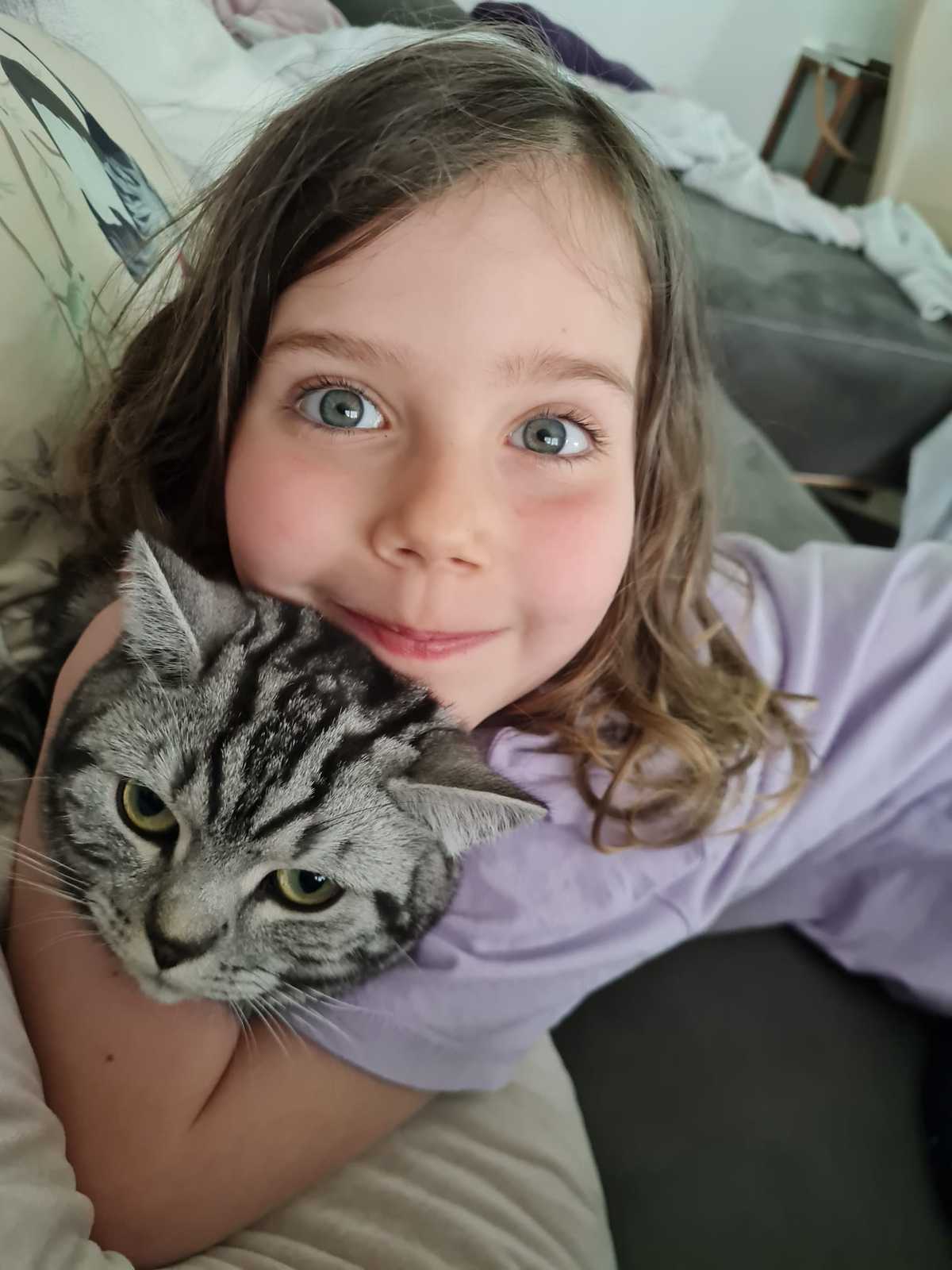 Ein Mädchen liegt auf einem Sofa und hält eine grau-schwarz gemusterte Katze im Arm. Beide blicken in die Kamera. Das Mädchen trägt ein hellviolettes Oberteil. Im Hintergrund sind ein graues Sofa, Kissen und Decken zu sehen.
