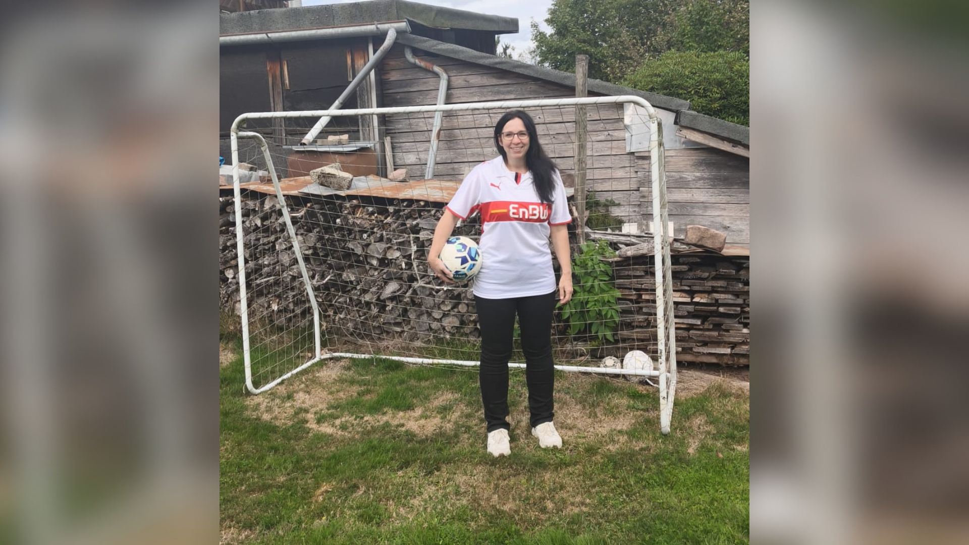 Eine Frau trägt ein weißes Trikot und hält einen Fußball im Arm während sie vor einem Tor steht.
