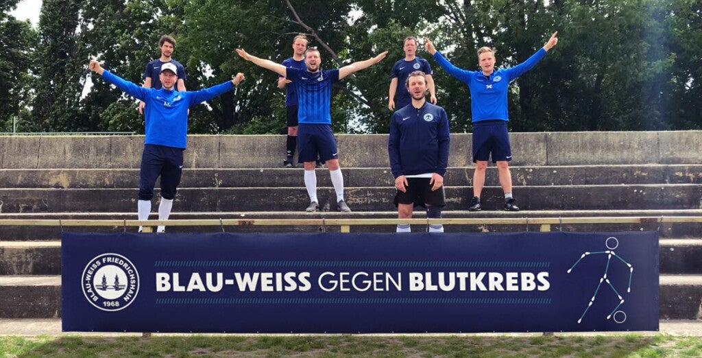Eine Gruppe von Männern in blauen Uniformen steht mit erhobenen Armen auf einer Treppe. Darunter ist ein Banner mit dem Text "Blau-Weiss Gegen Blutkrebs"