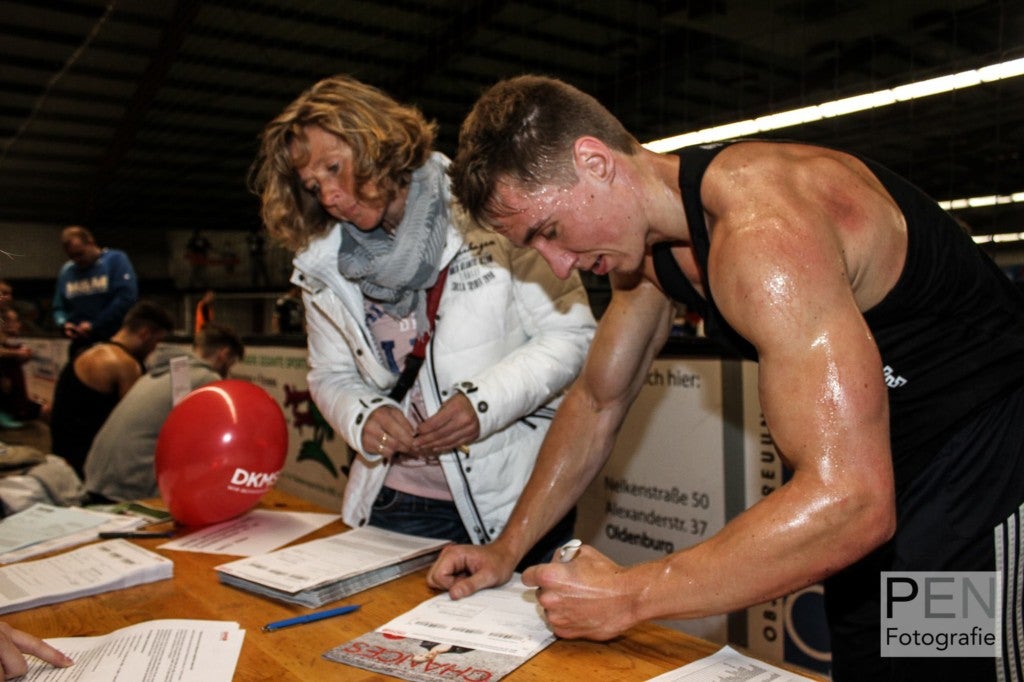 Ein Mann signiert etwas. Er trägt ein schwarzes Trägerhemd