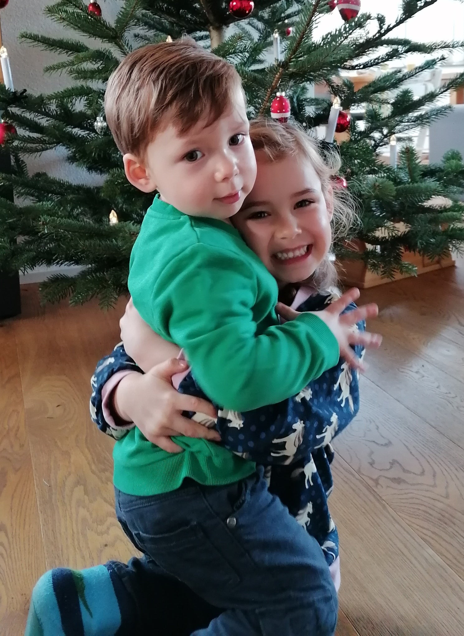 zwei kleine Kinder umarmen sich vor einem Weihnachtsbaum