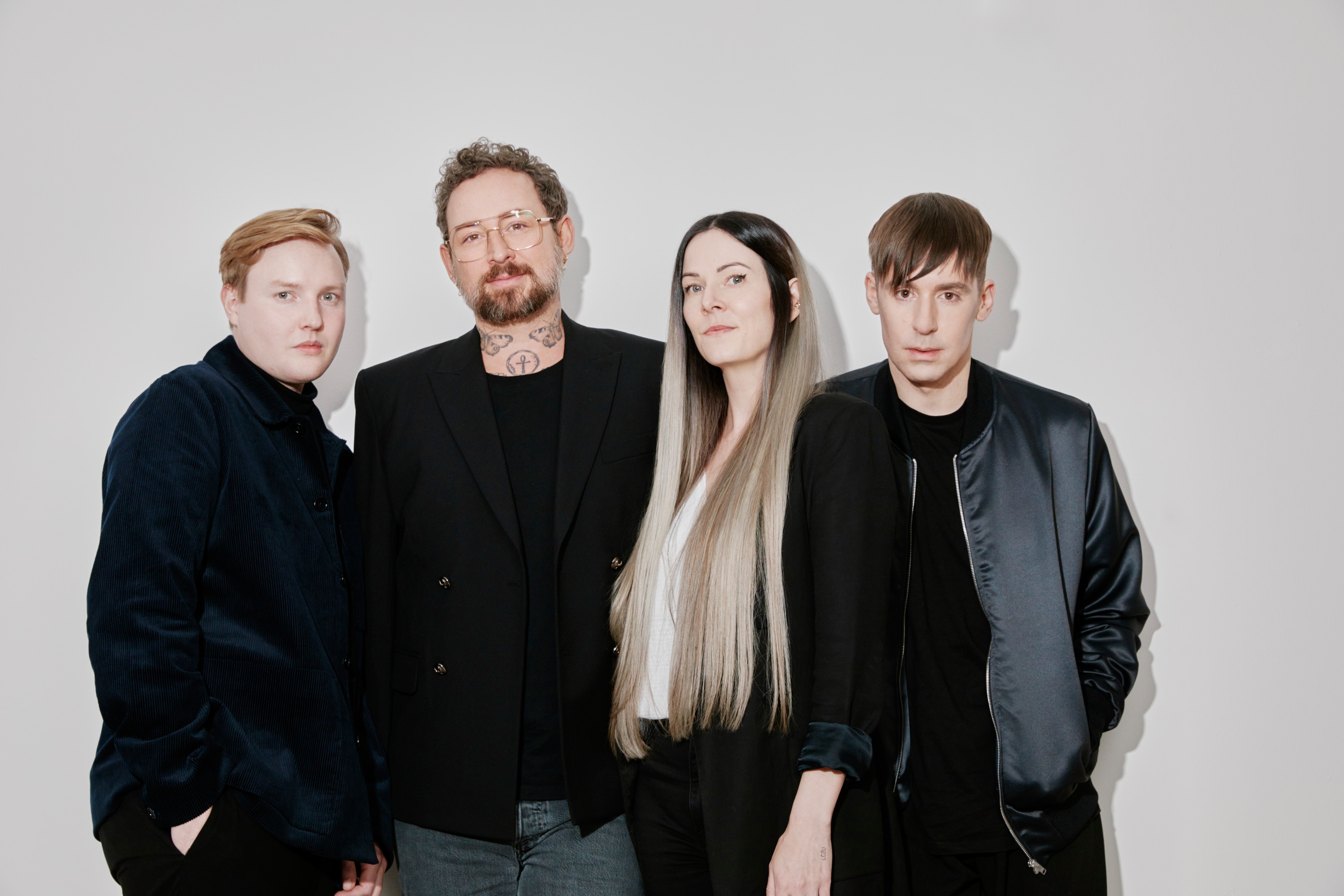 Vier Personen stehen nebeneinander vor einem weißen Hintergrund. Zwei Männer tragen dunkle Jacken oder Sakkos, eine Frau mit langen, glatt getragenen Haaren steht zwischen ihnen und trägt ein schwarzes Outfit. Alle blicken in die Kamera. Die Kleidung ist überwiegend in dunklen Farben gehalten.
