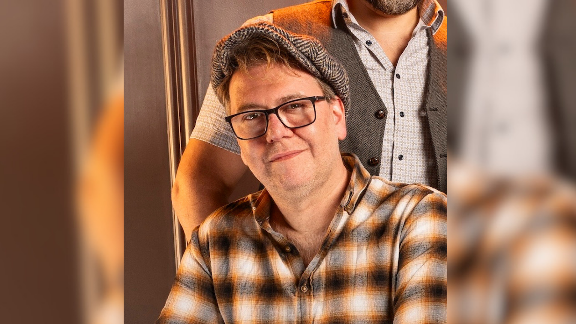 Ein Mann trägt ein kariertes Hemd, Brille und Schiebermütze. 