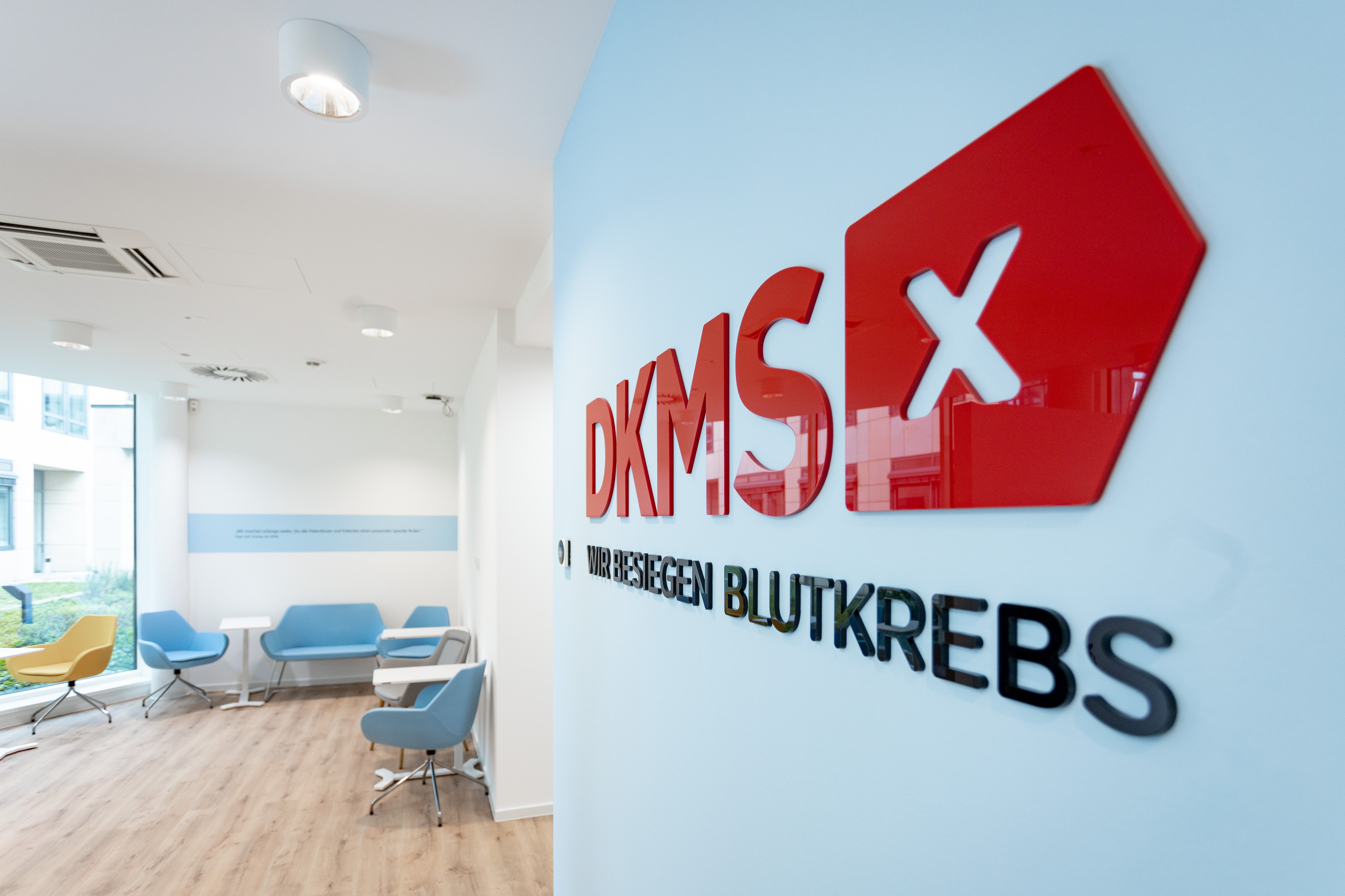 Innenansicht eines DKMS-Büros mit Logo an der Wand und Wartebereich mit Sesseln.