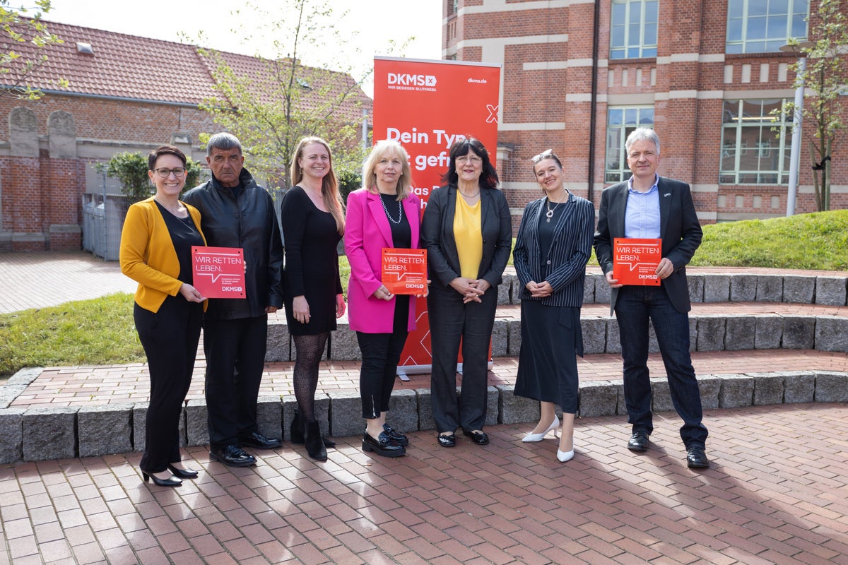 Sieben Personen stehen auf einem gepflasterten Platz vor einem roten DKMS-Aufsteller mit der Aufschrift „Dein Typ ist gefragt“. Einige von ihnen halten rote DKMS-Schilder mit der Botschaft „Wir retten Leben“. Im Hintergrund ist ein Backsteingebäude zu sehen. Die Gruppe wirkt engagiert und freundlich.