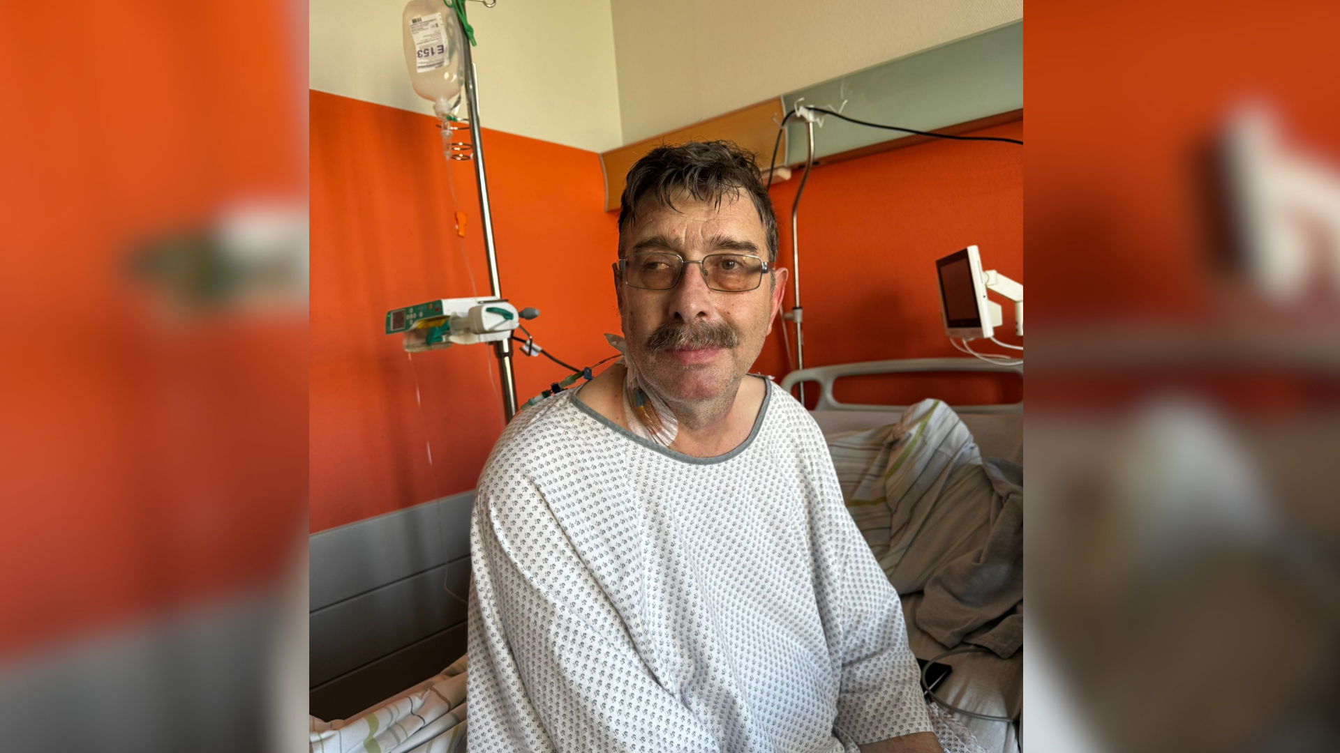 Ein Mann sitzt im Krankenkittel auf dem Rand eines Krankenhausbettes und ist am Hals an einen Tropf angebunden. 