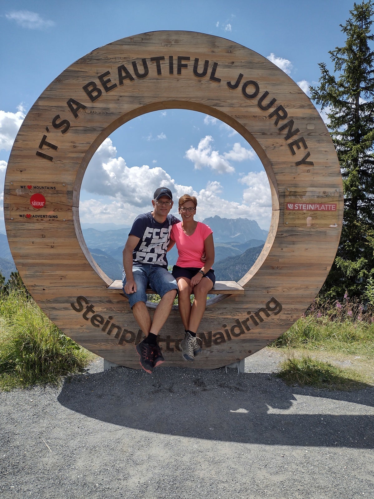 Ein Mann und eine Frau sitzen auf einem hölzernen Rundrahmen mit der Aufschrift „IT'S A BEAUTIFUL JOURNEY – Steinplatte Waidring“. Beide tragen sportliche Kleidung und Wanderschuhe. Im Hintergrund sind Berge, Wolken und blauer Himmel zu sehen. Der Rahmen steht an einem Aussichtspunkt auf einem Wanderweg.
