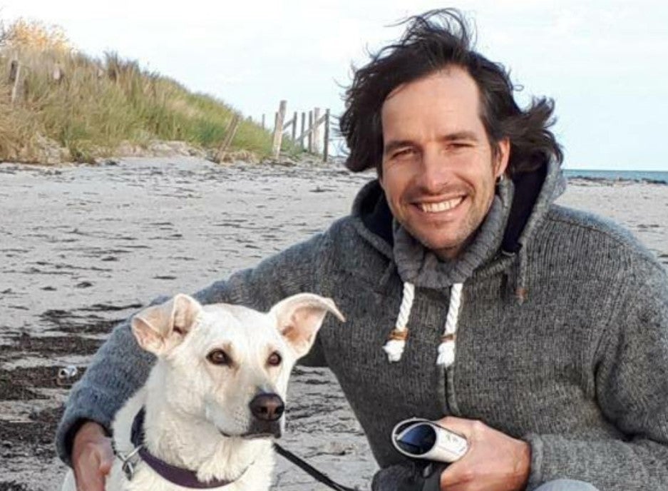 Ein Mann mit dunklem Haar und grauem Kapuzenpullover kniet am Strand neben einem weißen Hund mit Leine. Beide blicken in die Kamera. Der Mann hält eine Kamera in der Hand und lächelt. Im Hintergrund sind Sand, Dünengras und das Meer zu sehen.
