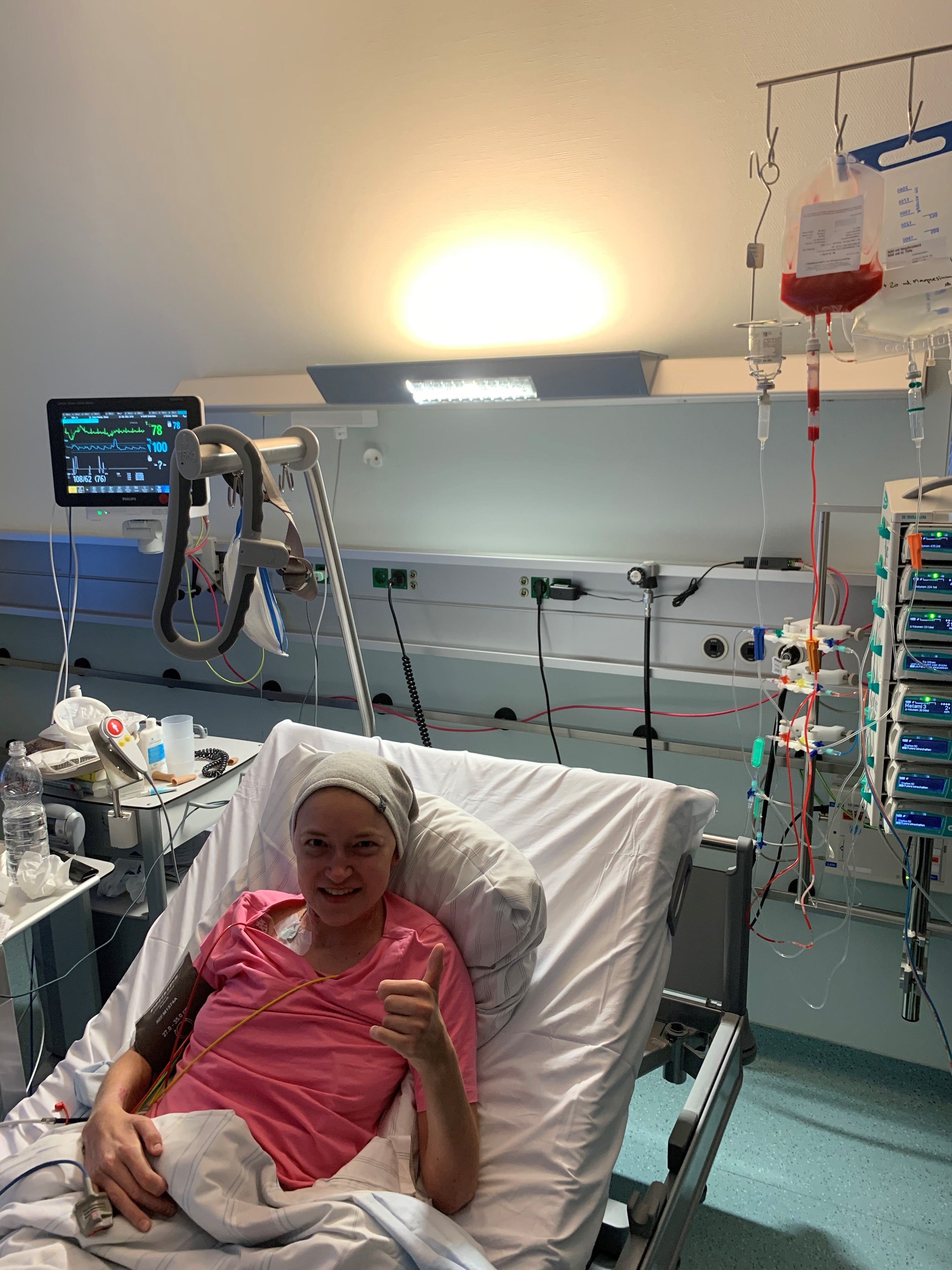 Eine Frau liegt in einem Krankenhausbett und zeigt mit einem Lächeln den Daumen nach oben. Sie trägt ein rosafarbenes Shirt und ein helles Tuch auf dem Kopf. Neben ihr sind medizinische Geräte und Infusionen zu sehen, darunter ein Beutel mit roten Stammzellen.