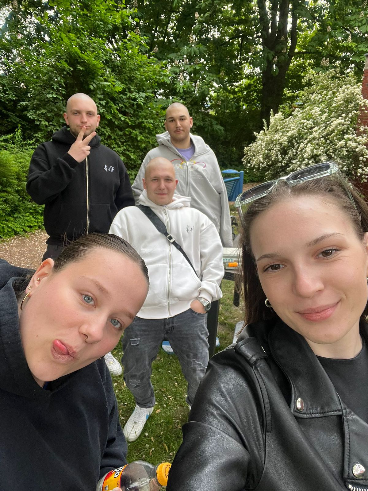 Drei Männer und zwei Frauen stehen im Freien vor Bäumen und Sträuchern. Eine Frau im Vordergrund hält das Handy für ein Selfie. Zwei Personen tragen weiße Oberteile, drei Personen schwarze. Im Hintergrund sind ein blauer Mülleimer und ein Anhänger zu sehen. Alle Personen schauen in die Kamera.