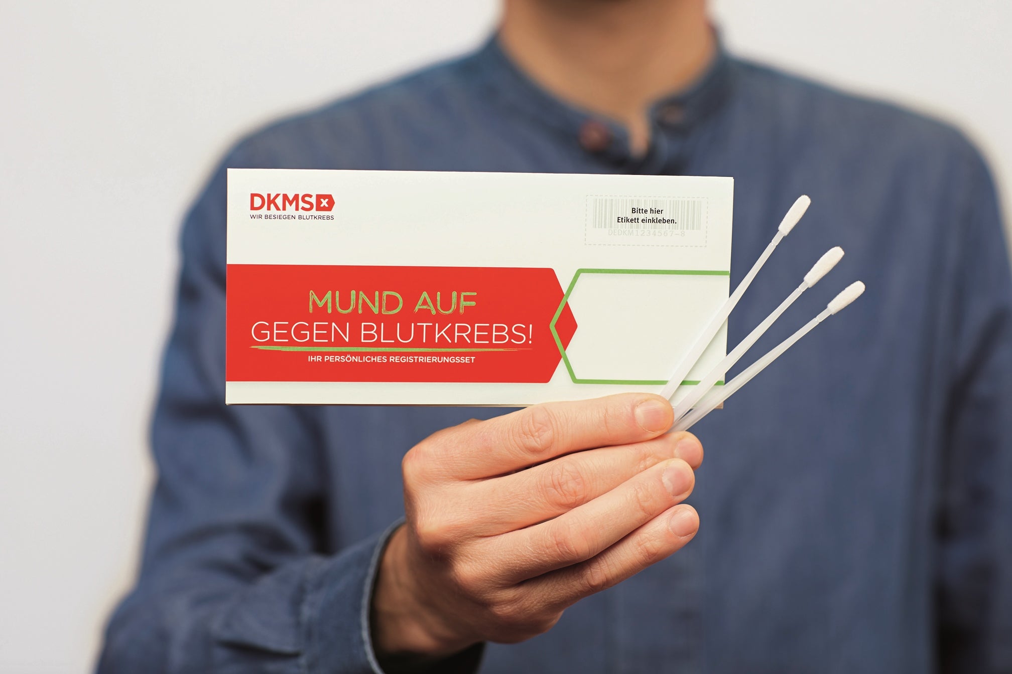 Eine Person in blauer Kleidung hält das DKMS-Registrierungsset mit der Aufschrift „Mund auf gegen Blutkrebs!“ sowie drei Wangenabstrichstäbchen in der Hand 