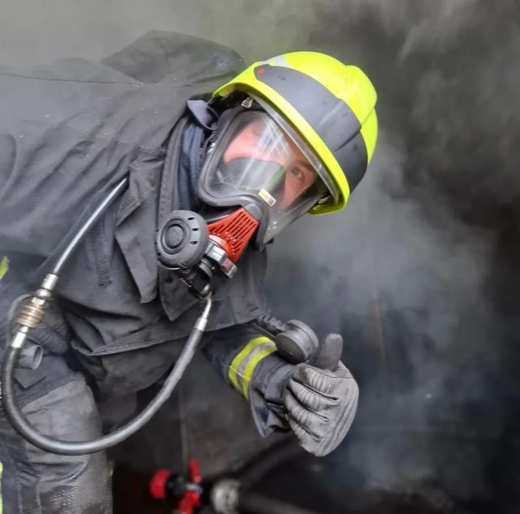 Eine Feuerwehrkraft in kompletter Schutzausrüstung mit Atemschutzmaske und gelbem Helm befindet sich in einem verrauchten Umfeld. Die Person beugt sich leicht nach vorne und zeigt den Daumen nach oben. Im Hintergrund sind Schläuche und ein Strahlrohr erkennbar.
