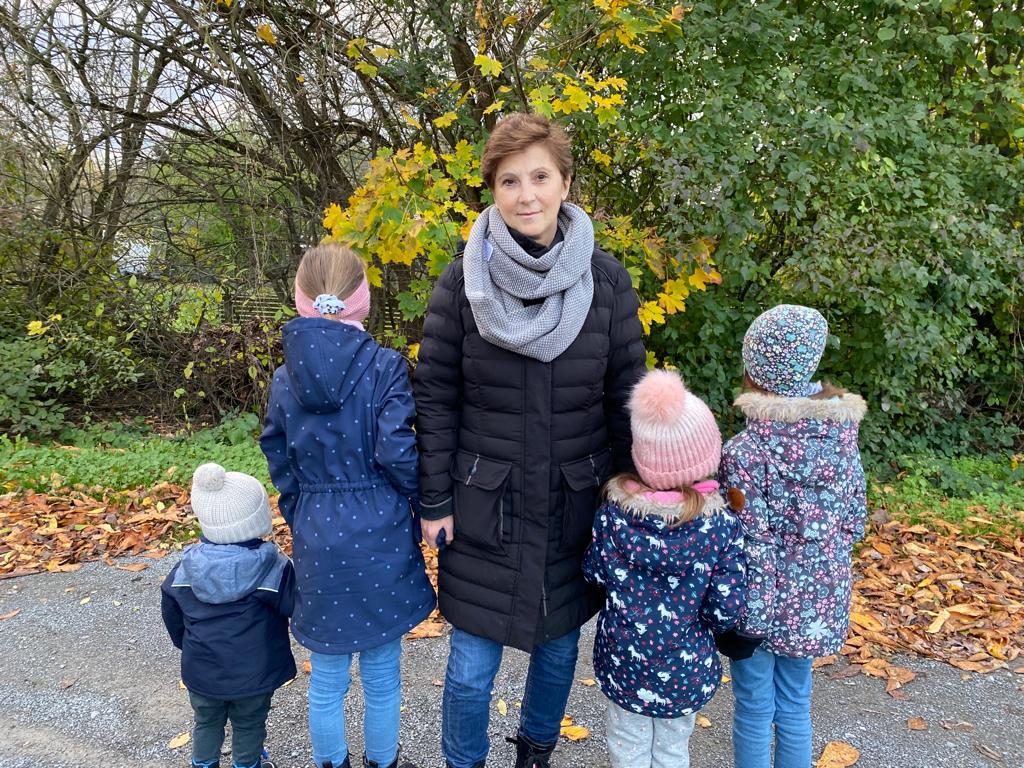 Eine Frau steht auf einem herbstlich mit Laub bedeckten Weg, umgeben von vier Kindern. Sie trägt eine schwarze Steppjacke, einen grauen Schal und blickt in die Kamera. Die Kinder stehen mit dem Rücken zur Kamera und tragen bunte Winterjacken und Mützen. Im Hintergrund sind Bäume mit herbstlich gefärbtem Laub zu sehen.