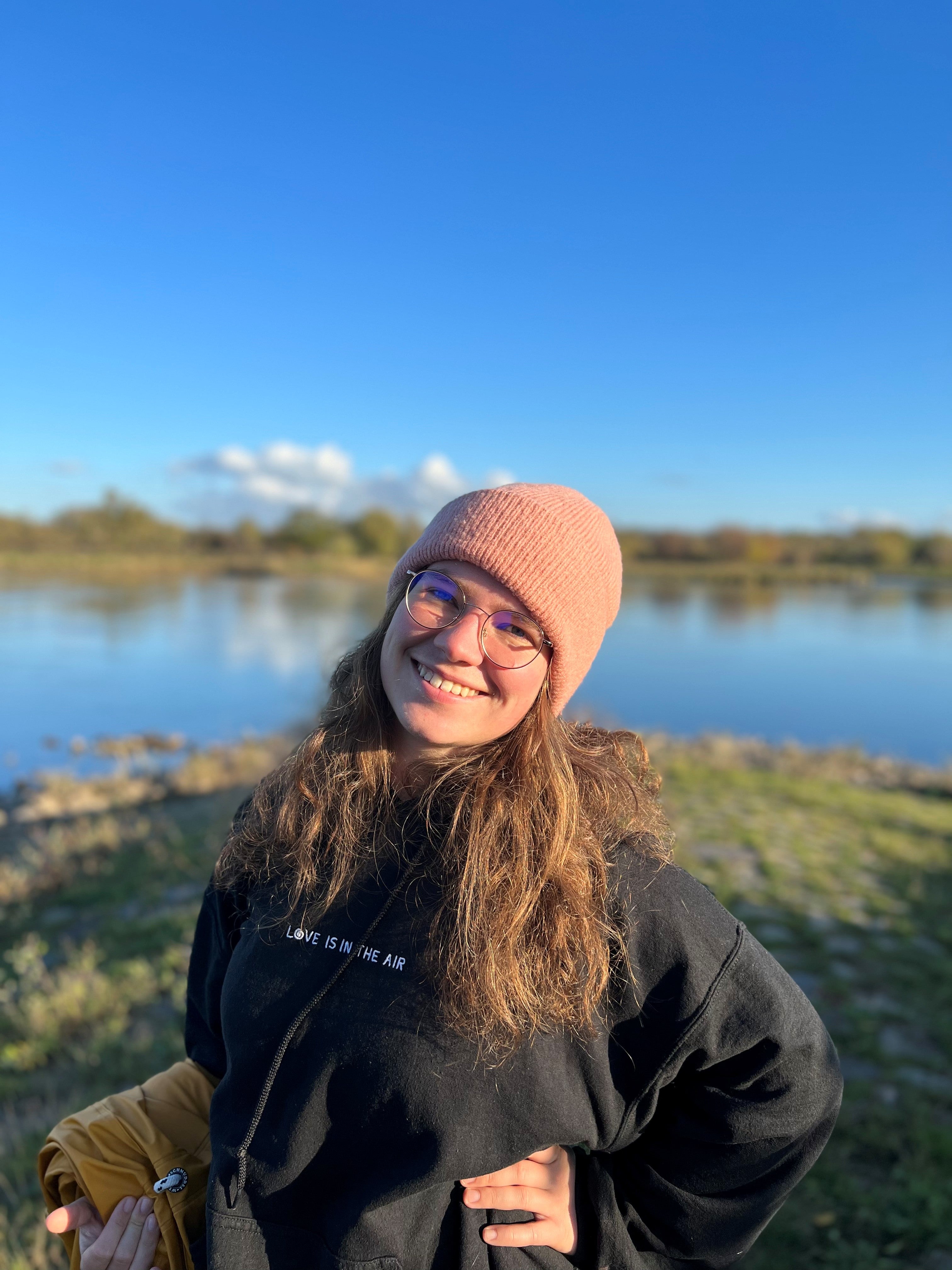 Fabienne steht draußen vor einem See und lächelt in die Kamera. Sie trägt einen schwarzen Hoodie und eine rosa Mütze. 