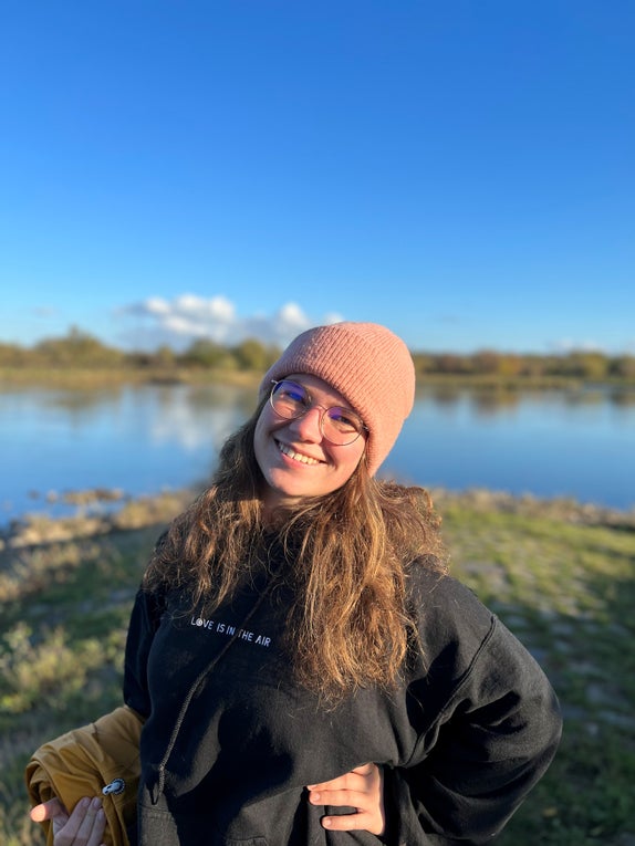 Fabienne steht draußen vor einem See und lächelt in die Kamera. Sie trägt einen schwarzen Hoodie und eine rosa Mütze. 