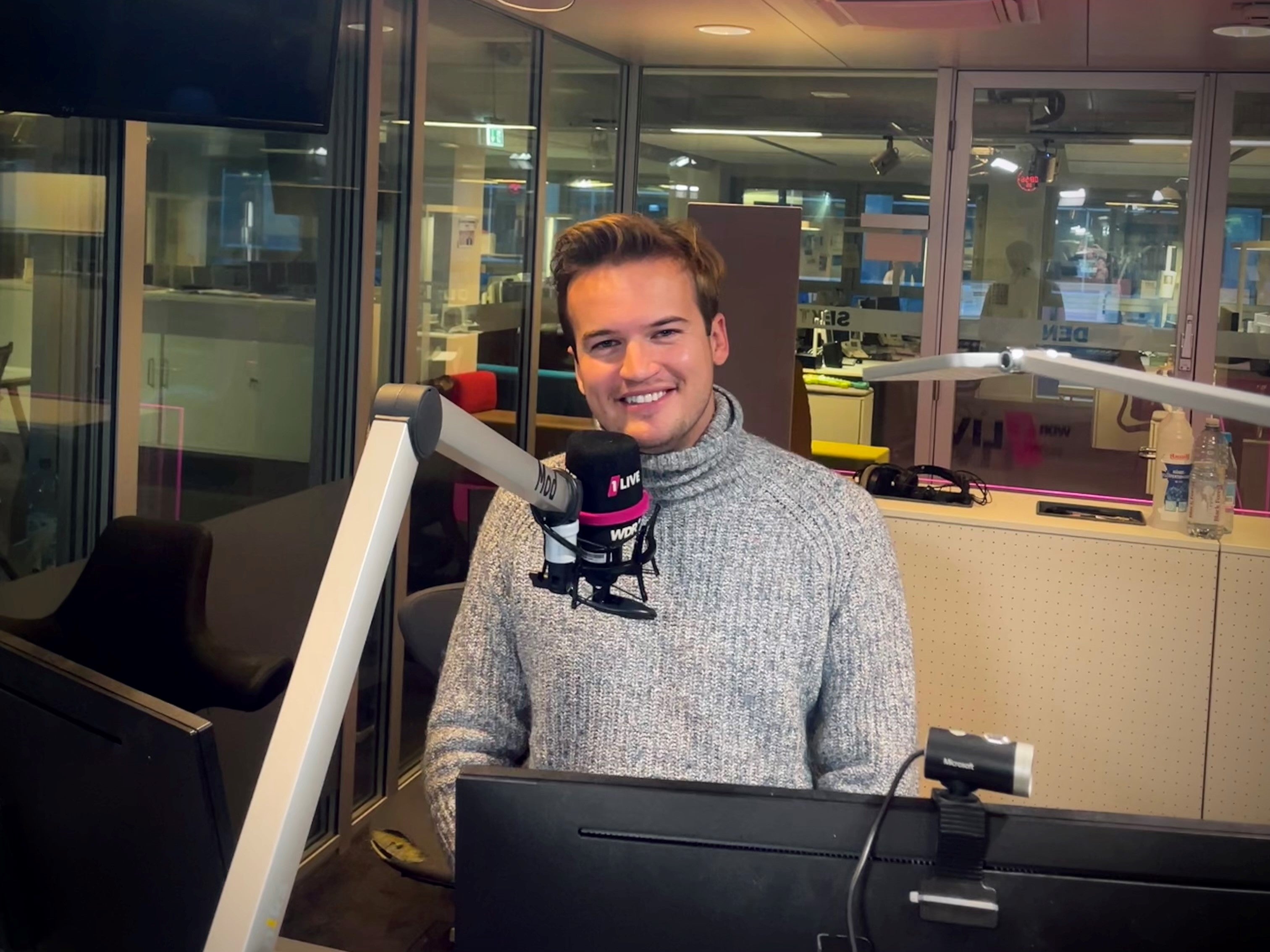 ein Mann sitzt hinter einem 1Live Mikrofon und lächelt in die Kamera
