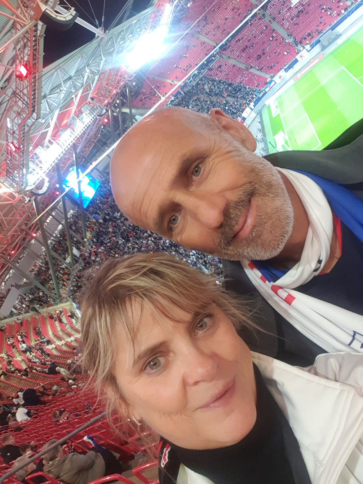 Ein Mann und eine Frau machen ein Selfie im Stadion. Beide tragen helle Jacken und lächeln in die Kamera. Im Hintergrund sind rote Sitzreihen, ein beleuchtetes Tribünendach und ein Fußballfeld mit laufendem Spiel zu sehen.