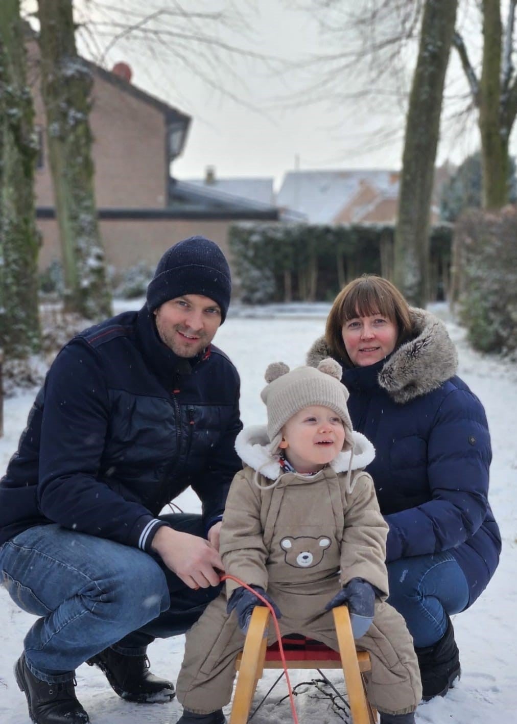 Ein Mann, eine Frau knien neben einem Kind, das auf einem Schlitten im Schnee sitzt.