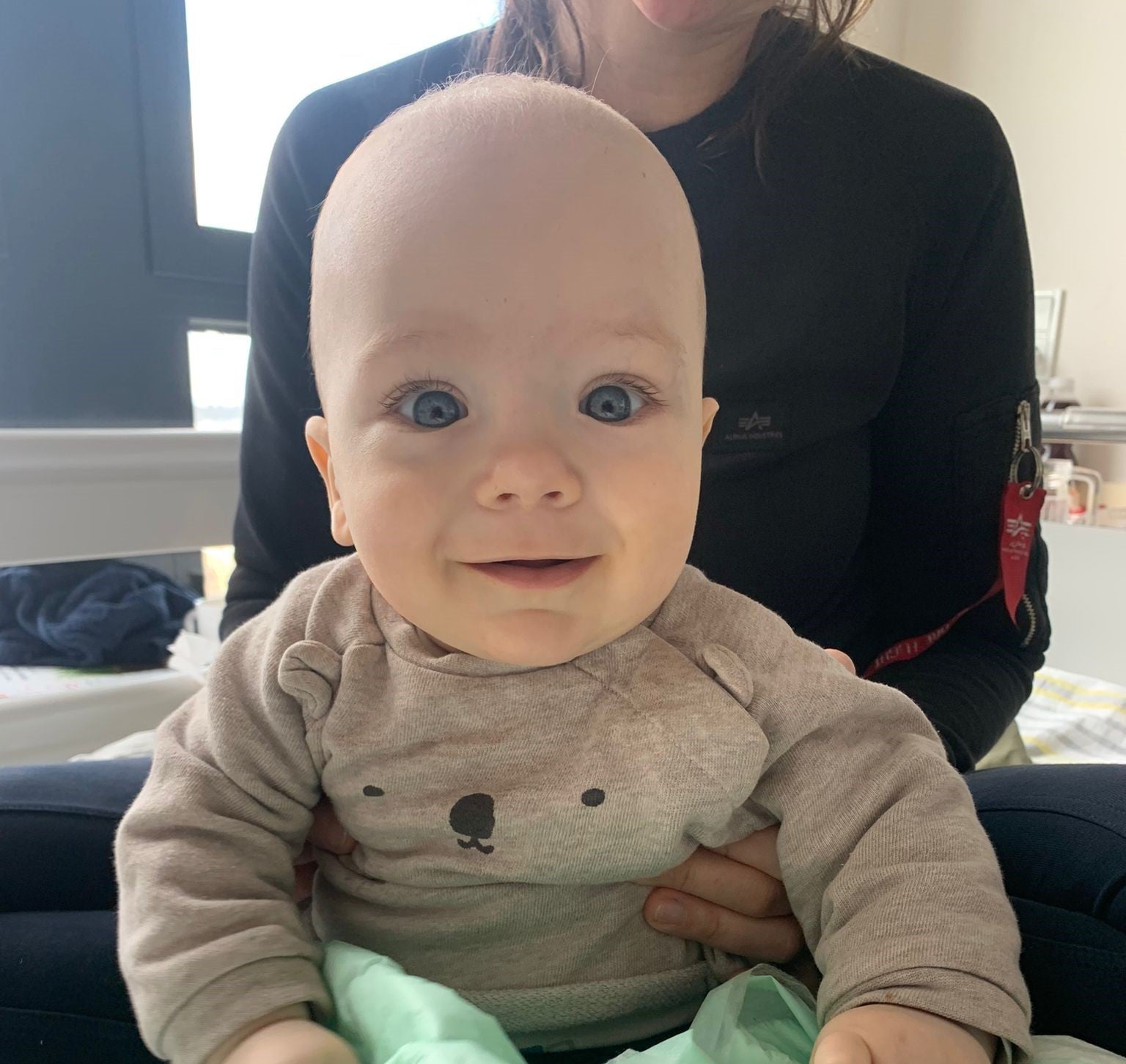 Ein Baby mit großen blauen Augen und einem beigefarbenen Pullover mit Bärengesicht sitzt auf dem Schoß einer Frau. Das Baby schaut lächelnd in die Kamera. Im Hintergrund ist ein Fenster und ein helles Zimmer zu sehen.