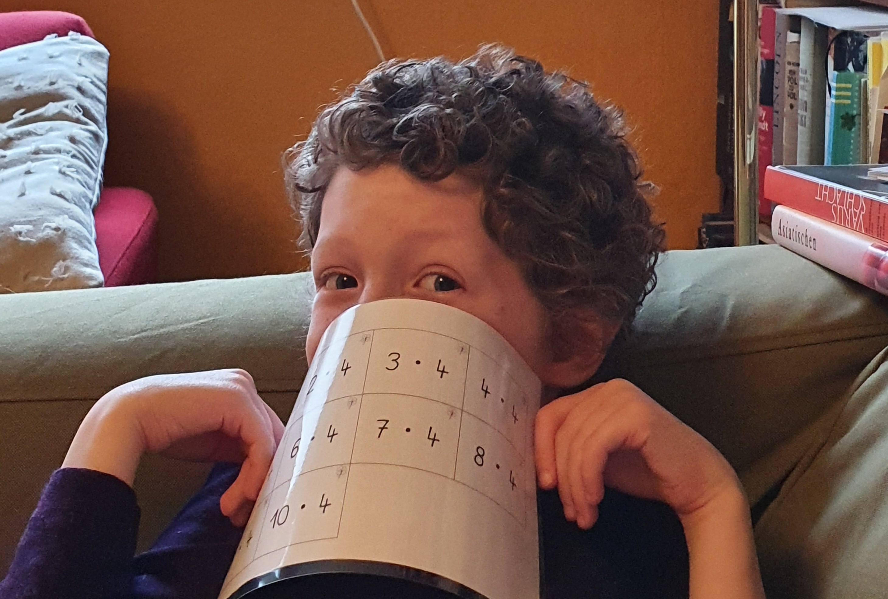 Ein Kind mit lockigen Haaren sitzt auf einem Sofa und hält sich ein Blatt Papier mit Rechenaufgaben spielerisch vor den Mund. Das Kind blickt mit einem verschmitzten Ausdruck zur Seite. Im Hintergrund sind ein rotes Kissen, Bücherregale und ein orangefarbener Wandbereich zu sehen.