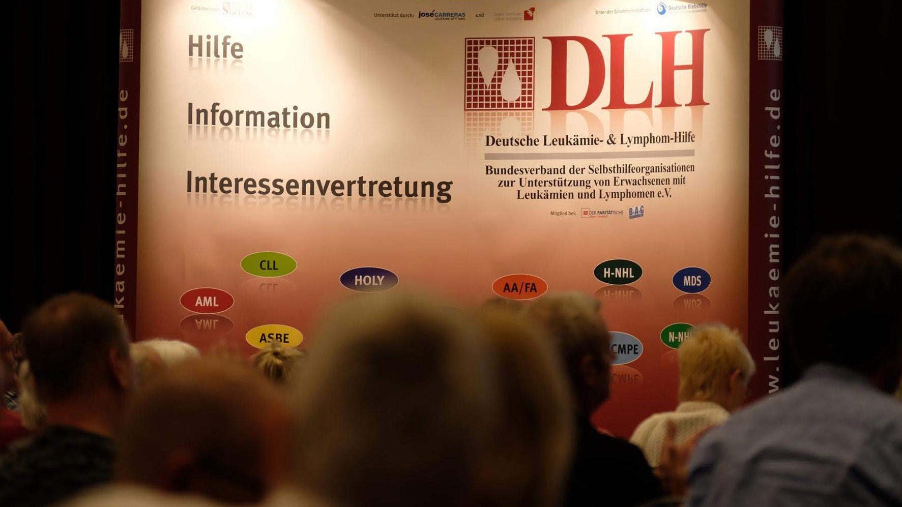 Eine Gruppe von Menschen vor einem Plakat. Auf der linken Seite die Worte "Hilfe", Information, "Interessenvertretung". Auf der rechten Seite ein Logo Deutschen Leukämie- & Lymphom-Hilfe e.V. 