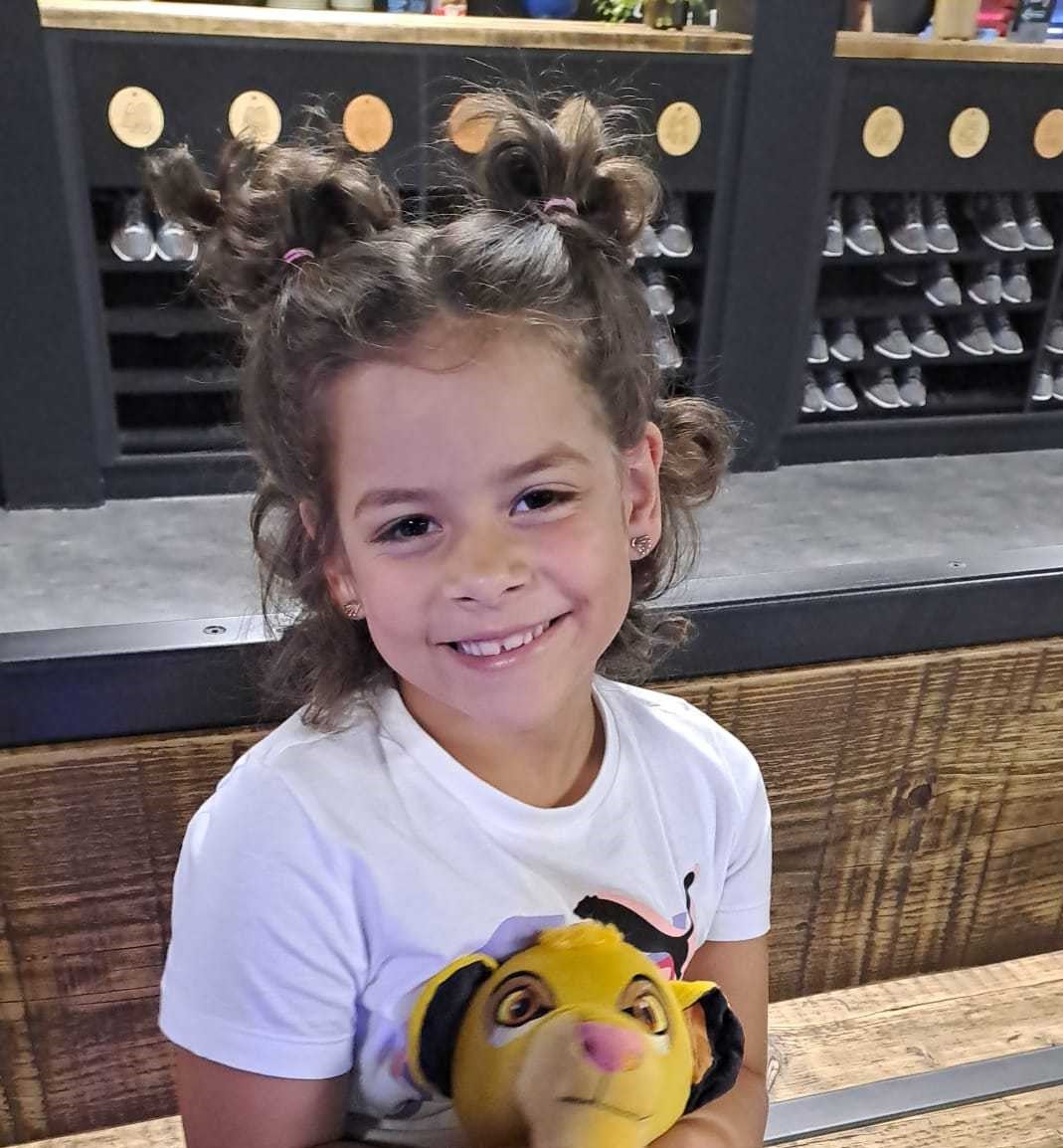 Ein kleines Mädchen mit lockigem Haar und mehreren Zöpfen sitzt auf einer Bank. Sie trägt ein weißes T-Shirt und hält ein Plüschtier aus dem Film „Der König der Löwen“ im Arm. Im Hintergrund ist ein Regal mit zahlreichen Paar Schuhen zu sehen.