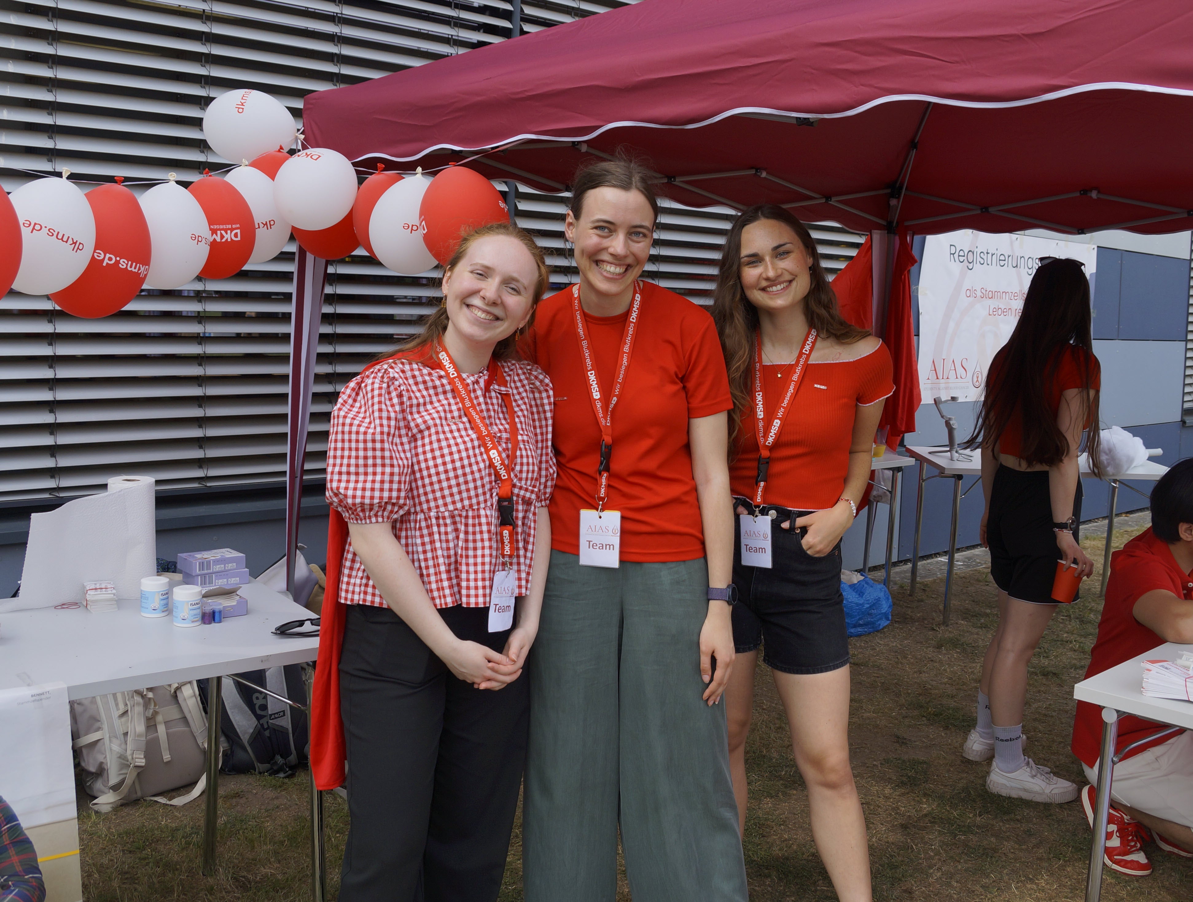 Drei Personen vor einem Stand mit Pavillons, Luftballons und Tischen