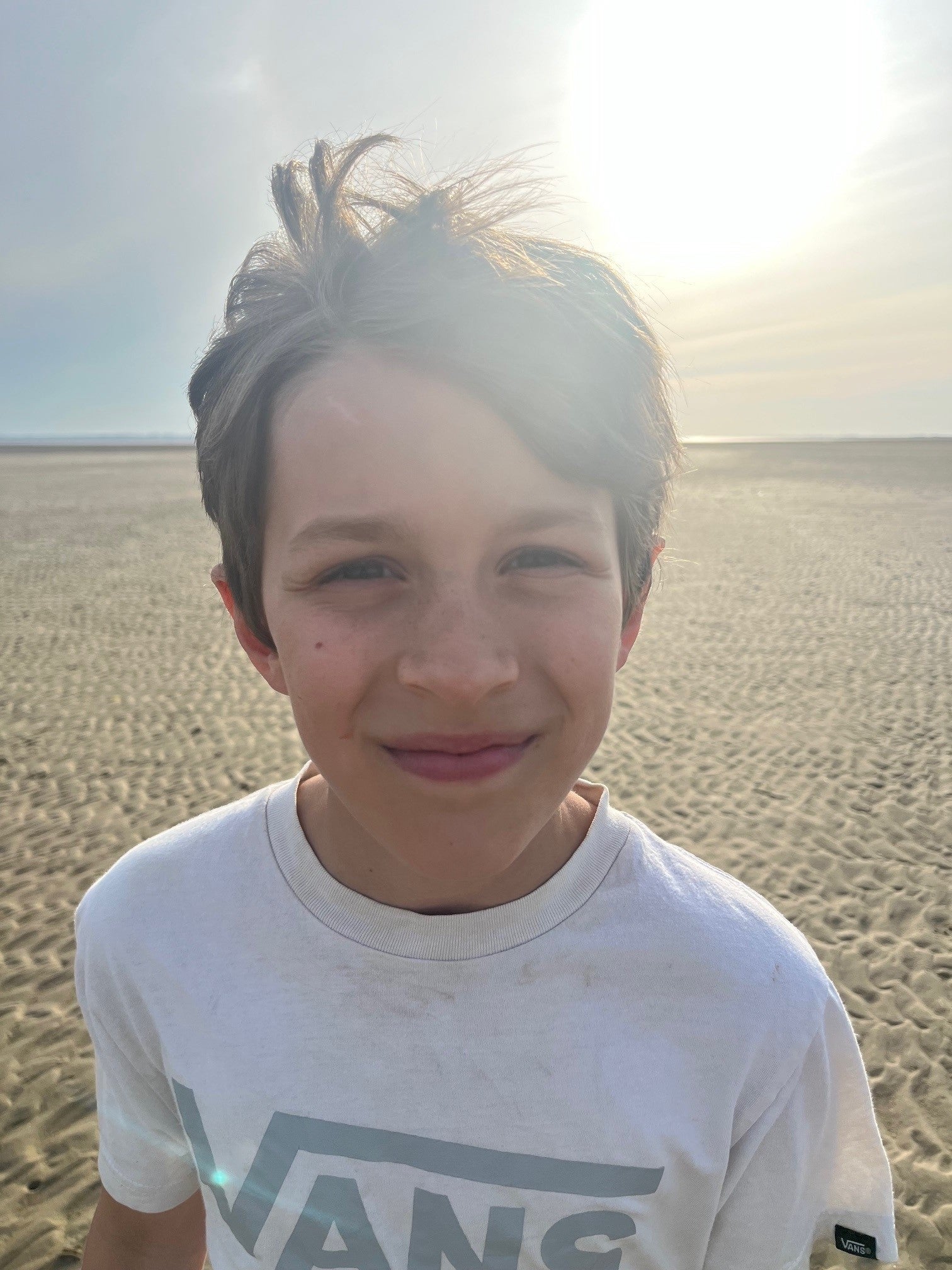 Ein Junge mit braunen Haaren und weißem T-Shirt lächelt in die Kamera. Hinter ihm ist die Sonne und ein Strand zu sehen.