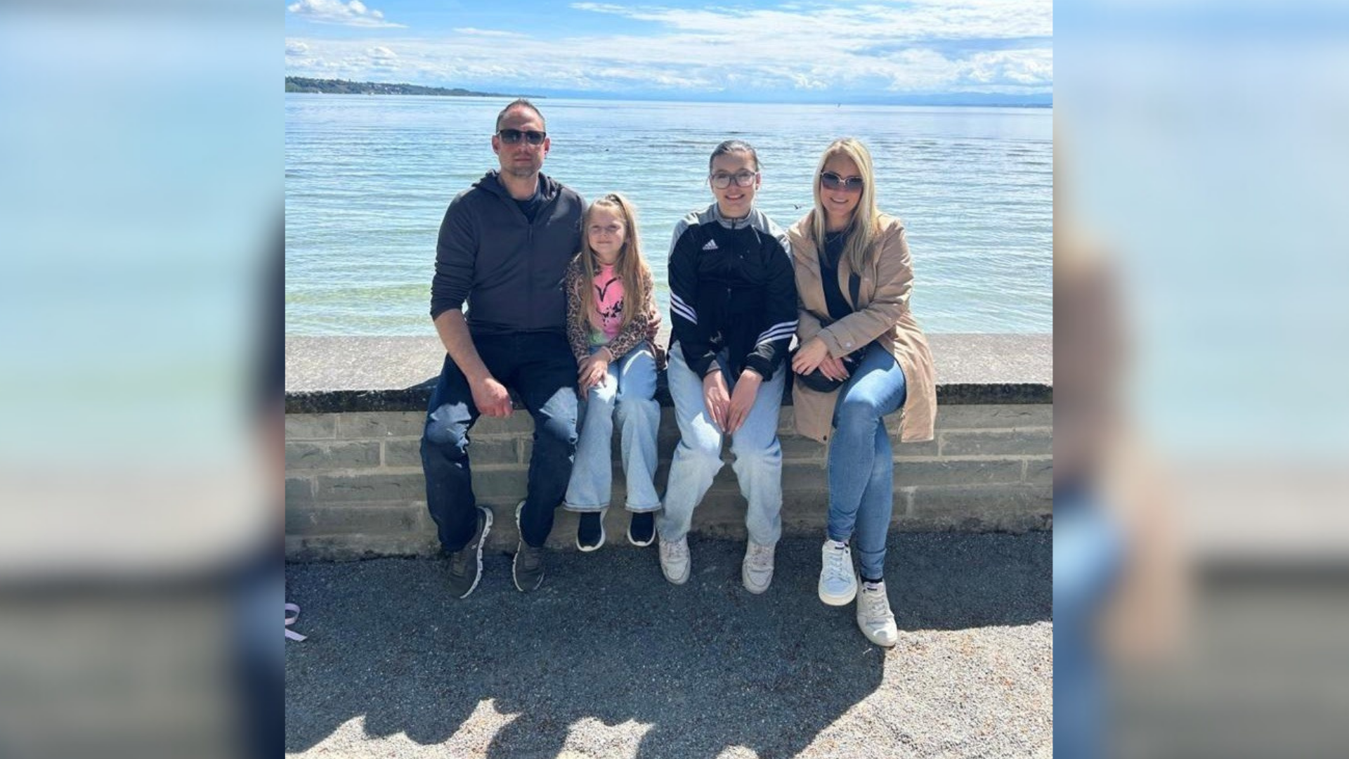 Ein Mann, zwei Mädchen und eine Frau sitzen auf einer Mauer am Ufer eines Gewässers. 