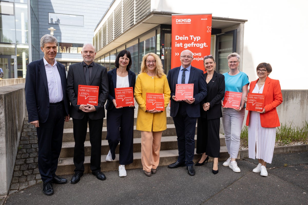 Acht Personen stehen vor einem modernen Gebäude und halten rote DKMS-Schilder mit der Aufschrift „Wir retten Leben“. Im Hintergrund steht ein DKMS-Aufsteller mit der Botschaft „Dein Typ ist gefragt“. Die Gruppe wirkt fröhlich und engagiert.